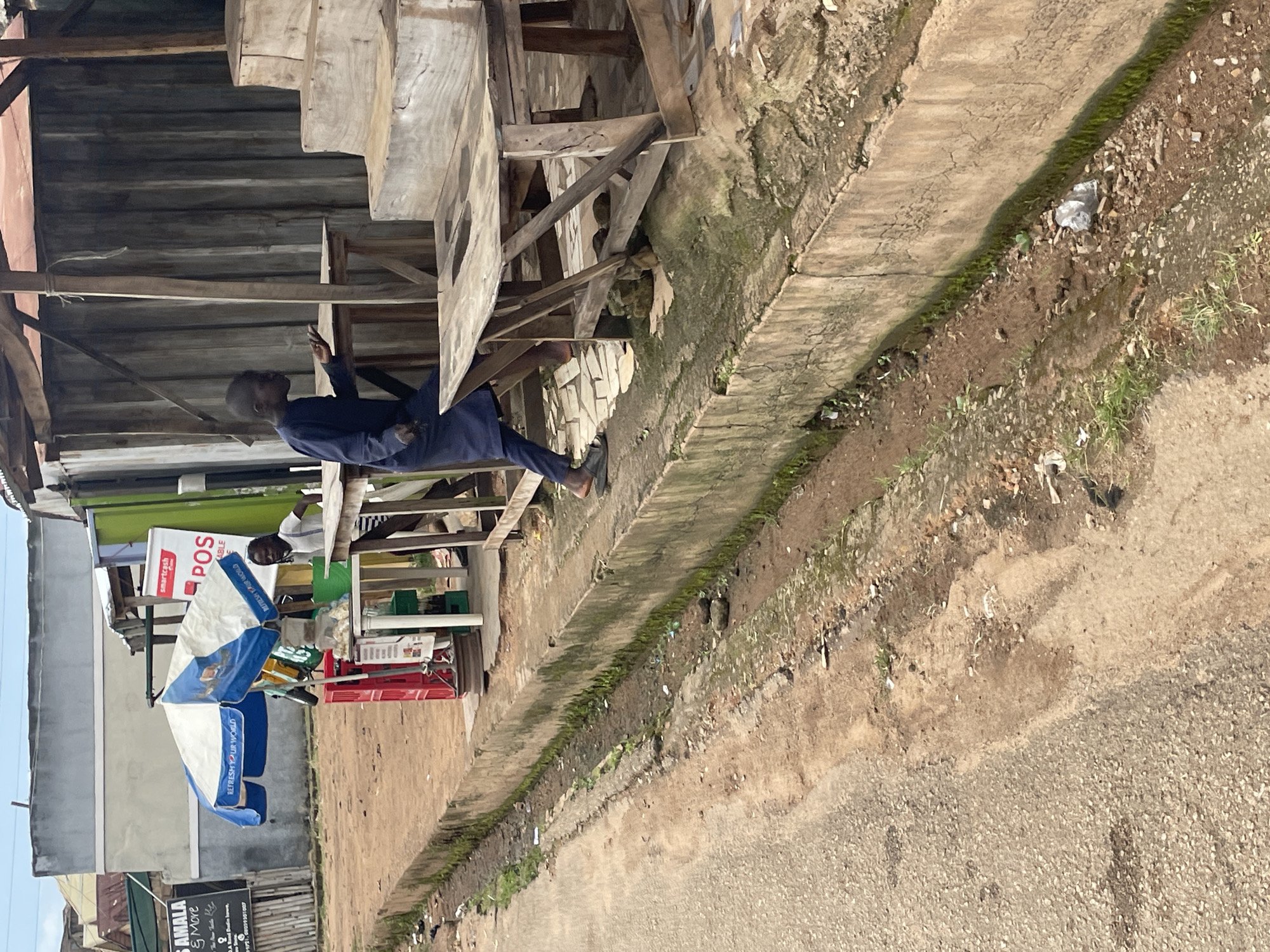 A boy, dressed in a dark blue tunic and trousers, stands on a paved surface with a broken drainage ditch running alongside it. He is facing to the right of the frame, with his left arm raised and palm open, as if gesturing or in conversation. He wears dark sandals. The boy is positioned near a series of wooden tables and tiered wooden platforms, which appear to be part of an outdoor market stall. In the background, a white and blue umbrella is set up, with a red plastic crate beneath it. A white sign with red lettering that says "POS" is visible behind the umbrella. Further in the background, a building with a gray facade and a dark, ribbed wall structure are visible. The ground is a mix of dirt, gravel, and patchy vegetation. The scene appears to be an outdoor market area in Jos, Nigeria.