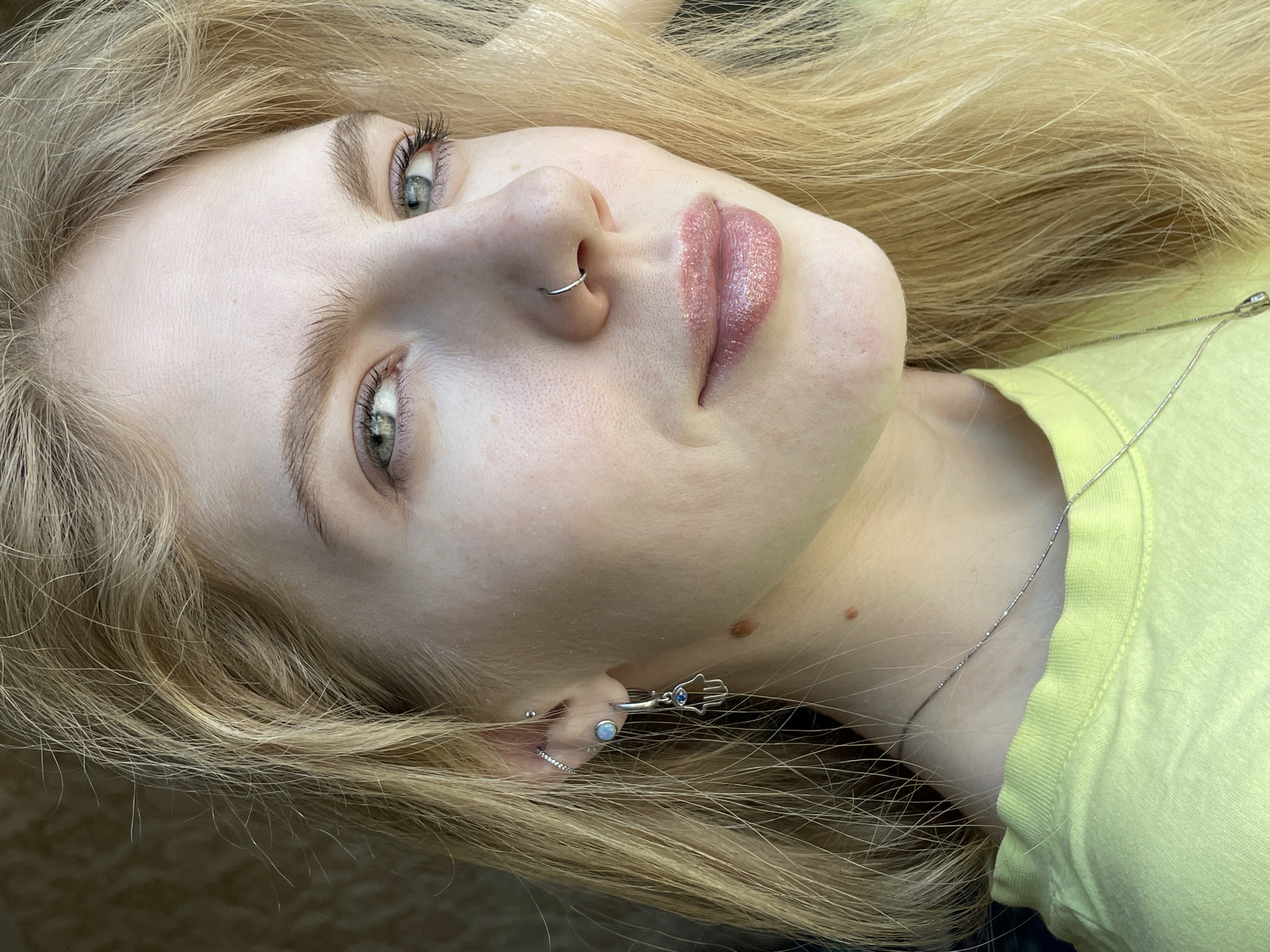 A close-up, slightly angled view of a fair-skinned person with long blonde hair. The person has light eyes and wears a silver nose ring. Their lips are adorned with a glossy lipstick or lip gloss. The person wears several earrings on one ear and a silver necklace. A light green or yellow-green garment is partially visible in the lower right. No information is provided that would suggest the image was taken in Izhevsk, Russia.