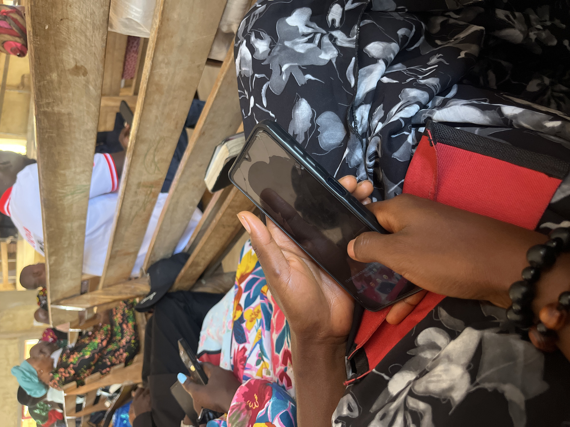 The image, taken in Bukuru, Nigeria, depicts a close-up view of a person holding a smartphone. The individual is seated and wearing a black and white floral print dress. Their hands are positioned around the phone, which displays a dark screen with some text and icons visible, including what appears to be a message interface. In the background, other people are seated on wooden benches, some of whom are blurred due to the shallow depth of field. The overall environment suggests an indoor gathering or waiting area, possibly a church or community hall, given the presence of wooden seating and the attire of some individuals. The lighting is relatively dim, with natural light filtering in from an unseen source.