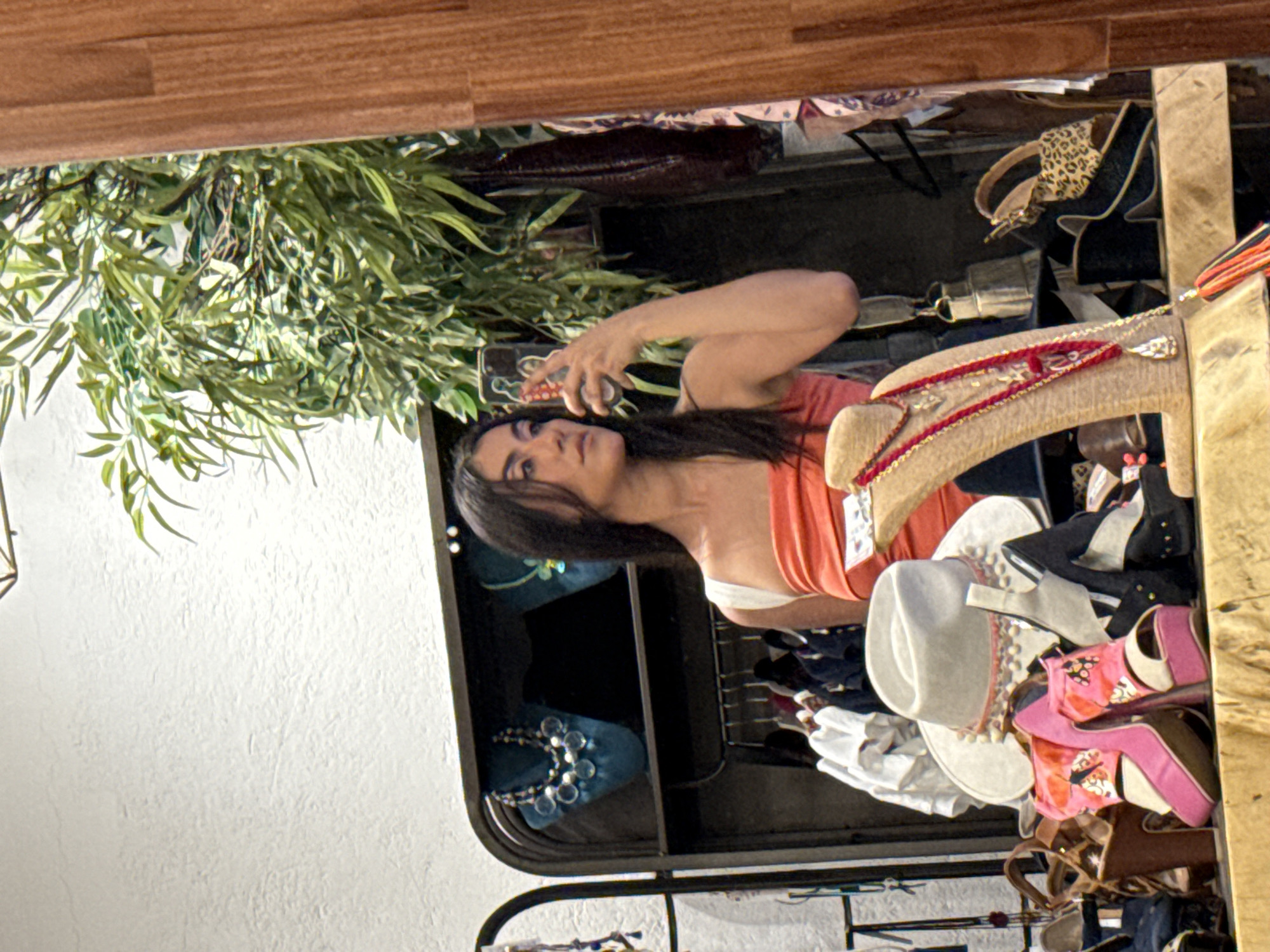 The image is a selfie of a woman in a Domingo Arenas, Mexico store, likely a boutique or shop specializing in accessories. The woman, with dark hair, is wearing an orange tank top. She's holding a smartphone up to take the photo, and her reflection is visible in a mirror.
Behind her is a display of various accessories. Several hats of different styles (white, straw, patterned) are stacked. Other items visible include shoes (pink heels, leopard-print flats) and jewelry. A faux plant is situated above the display, adding a decorative element to the scene. The setting appears to be inside a retail establishment, with clothing racks and other merchandise displayed. The overall impression is of a casual shopping environment.