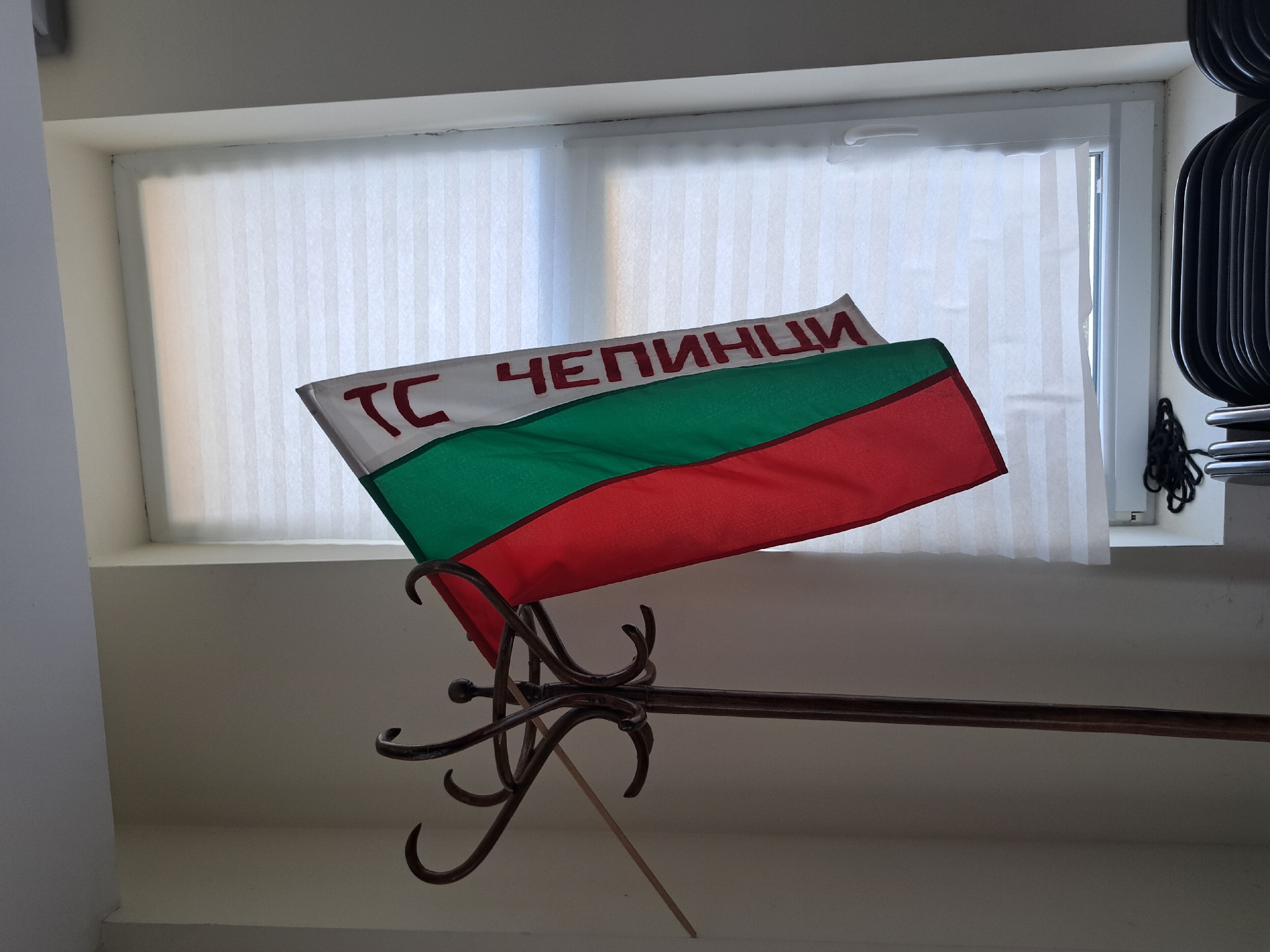 A flag with three horizontal stripes of white, green, and red is displayed. The white stripe at the top features red Cyrillic text that reads "ТС ЧЕПИНЦИ". The flag is attached to a decorative metal stand with multiple curved arms. The background shows a window covered with vertical blinds and a wall. There are also some black objects on the right side of the image that appear to be a wire rack or similar item. The lighting suggests it might be daytime, possibly indoors. The overall scene appears to be a display of a flag, possibly in a public space or a collection.