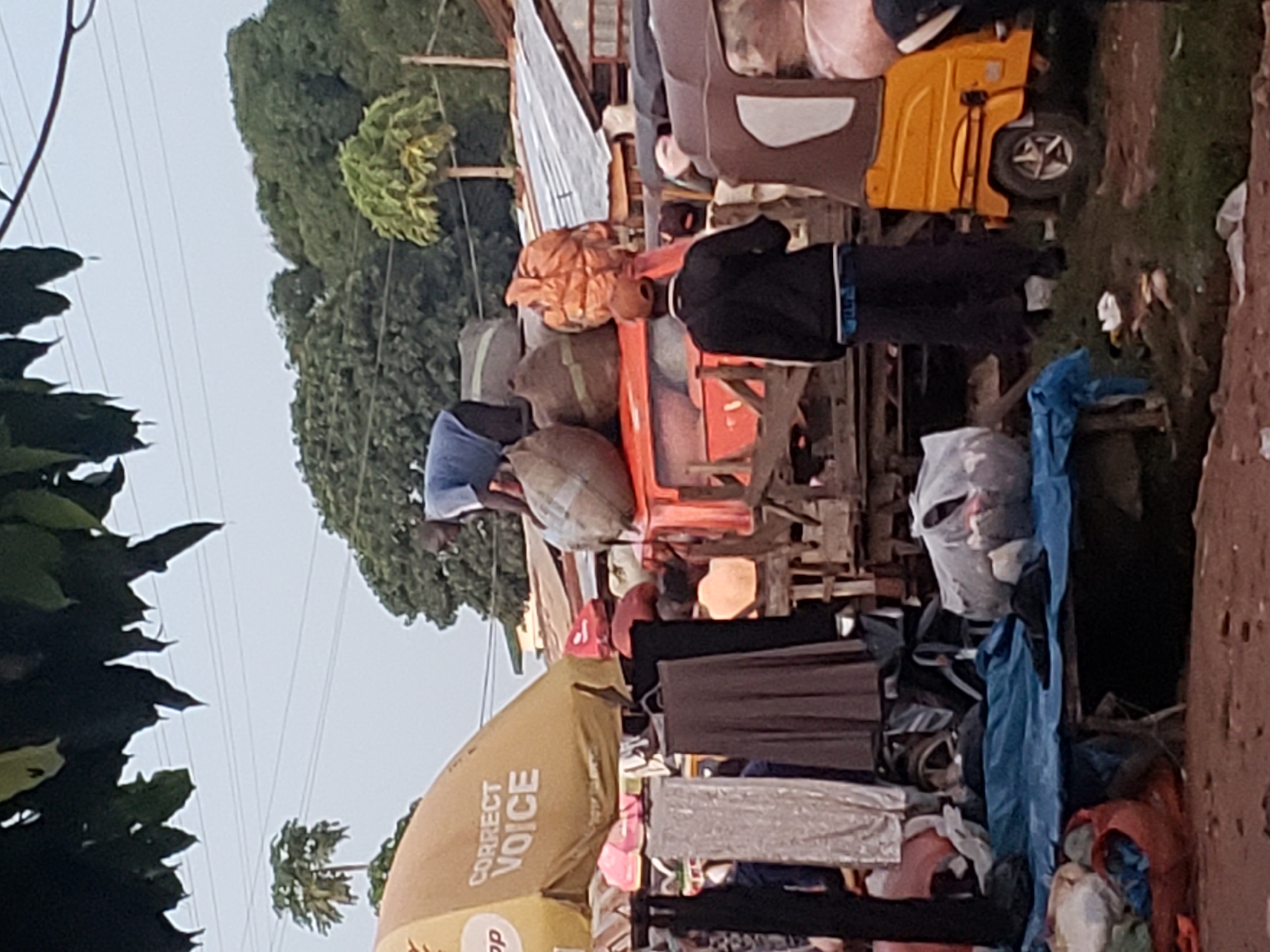 The image depicts a scene in a market in Kakuri Gwari, Nigeria. In the foreground, a blue tarp covers a collection of goods, including bags and bundles of clothing. To the left of the tarp, several pieces of clothing, including a black long-sleeved garment and a light-colored patterned garment, are hanging. Further to the right, a yellow and brown auto-rickshaw is parked. Behind the rickshaw, a man wearing a black jacket and dark pants stands facing away from the camera. In the middle ground, a red truck is partially visible, loaded with large sacks. A man in a blue shirt is standing on top of the sacks on the truck, appearing to be arranging them. Another man, wearing a brown cap and a black jacket, stands near the truck. In the background, there are structures with corrugated metal roofs and trees. Overhead, power lines are visible against a pale sky.