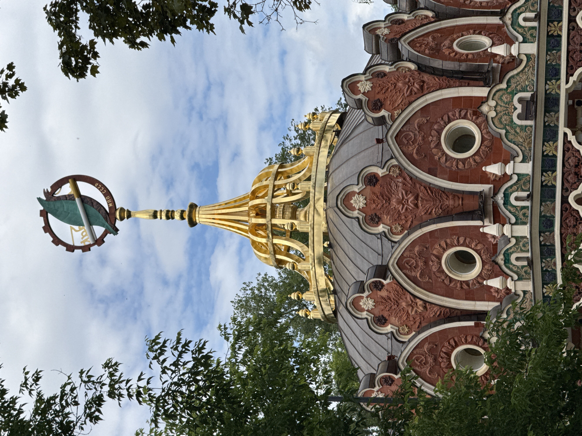 The image shows a close-up view of the roof and spire of the Main Pavilion of the All-Russia Exhibition Centre (VDNKh) in Moscow, Russia. The pavilion's dome is covered in dark material, likely metal, and features decorative red brickwork with white trim. Above the dome, a golden, ornate cupola rises, topped by a spire. The spire itself is crowned with a circular emblem containing a red gear, a green leaf, and a white cigarette, with the text "МОП СССР" visible. Surrounding the structure are lush green trees, with branches and leaves framing the top and sides of the image. The sky above is cloudy with patches of blue. The architectural style is characteristic of Soviet-era monumentalism, with elements of constructivism.