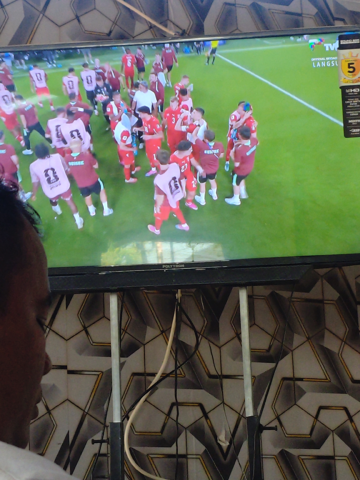 The image shows a television screen displaying a soccer match. The screen is positioned in a room with a patterned wallpaper. A person's head and shoulder are visible in the foreground, looking towards the television. On the screen, several soccer players in red and pink uniforms are on a green field. Some players are embracing, suggesting a celebration. A graphic on the top right corner of the screen shows "TV", "OFFICIAL BROADCAST", and "LANGSUNG" which translates to "Live" in Indonesian. Another graphic shows "5". The television appears to be a flat-screen model. The room is dimly lit, and the focus is on the television screen. There is no specific indication of the city of Parangsembilan, Indonesia, in the image itself.