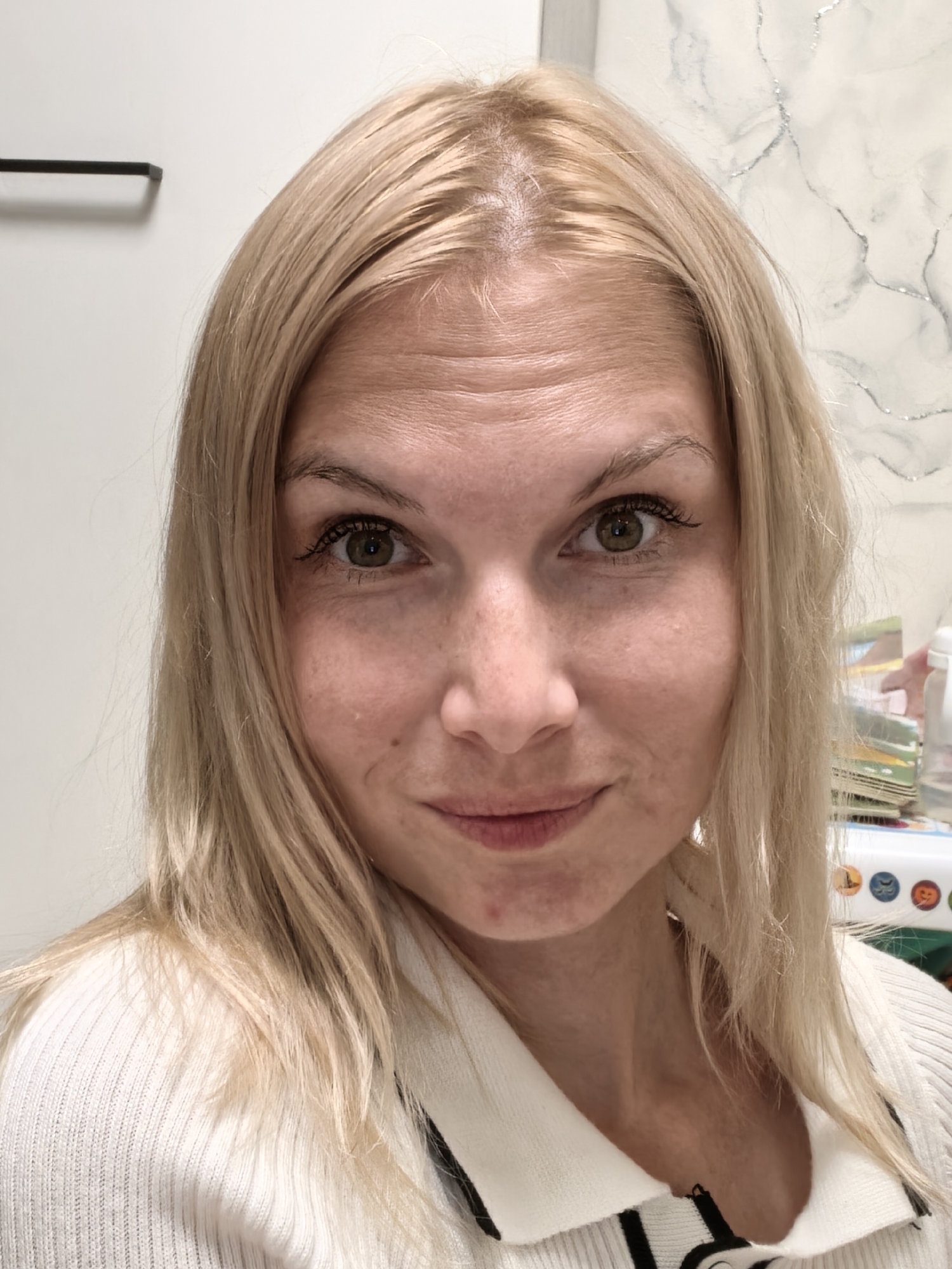 A blonde woman with green eyes, wearing a white sweater, is shown from the shoulders up. She has fair skin with freckles and appears to be looking directly at the camera with a slight smile. The background is a plain white wall with some decorative elements and objects on the right side. The image is a selfie taken indoors, with no discernible landmarks or context that would place it in Sterlitamak, Russia or any other specific city.
