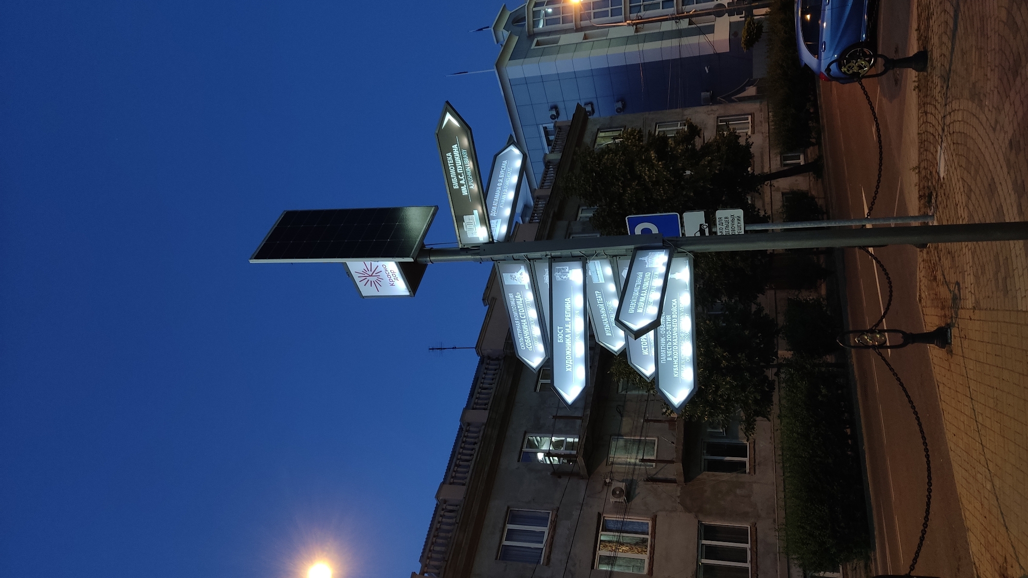 The image depicts a street signpost in Krasnodar, Russia, featuring multiple illuminated directional signs pointing to various cultural landmarks. The signs, written in Russian, include "СКУЛЬПТУРНАЯ КОМПОЗИЦИЯ «СОБАЧКИНА СТОЛИЦА»" (Sculptural Composition "Dog Capital"), "БЮСТ ХУДОЖНИКА И.Е. РЕПИНА" (Bust of Artist I.E. Repin), "МУЗЫКАЛЬНЫЙ ТЕАТР" (Musical Theater), "КРАЕВОЙ ХУДОЖЕСТВЕННЫЙ МУЗЕЙ ИМ. А.Ф. КОВАЛЕНКО" (Krasnodar Art Museum named after A.F. Kovalenko), and "ПАМЯТНИК-ОБЕЛИСК В ЧЕСТЬ 200-ЛЕТИЯ КУБАНСКОГО КАЗАЧЬЕГО ВОЙСКА" (Monument-Obelisk in Honor of the 200th Anniversary of the Kuban Cossack Army). Additionally, there are signs for "БИБЛИОТЕКА ИМ. А.С. ПУШКИНА" (Pushkin Library) and "ДОМ АТАМАНА Ф.Я. БУРСАКА" (Ataman F.Ya. Bursak's House). At the top of the signpost, a solar panel is visible, suggesting it powers the illuminated signs. The background features multi-story buildings with classical architectural elements and modern facades, as well as trees and a street with a blue car parked on the right. The sky is a deep blue, indicating twilight or early evening. A chain fence with decorative posts runs along the sidewalk.