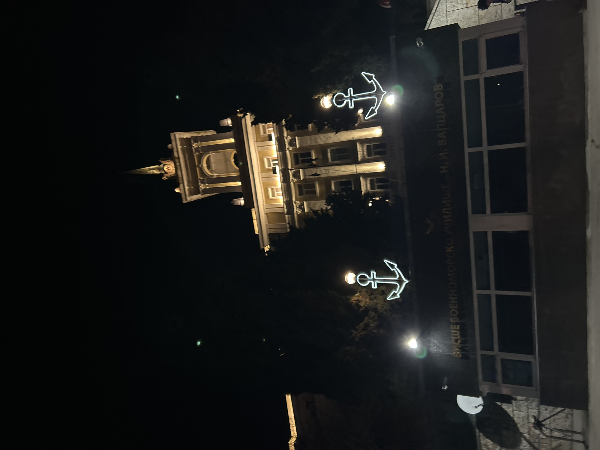 At night in Varna, Bulgaria, an outdoor shot shows the facade of a building and a tower illuminated with artificial lighting. The building, identified by a vertically-oriented plaque as “ВИСШЕ ВОЕННОМОРСКО УЧИЛИЩЕ .Н.И. ВАПЦАРОВ,” features lit anchor symbols above the entryway. A tower stands in the background, also illuminated. A satellite dish is mounted on the side of the building. No people are clearly visible in the frame.