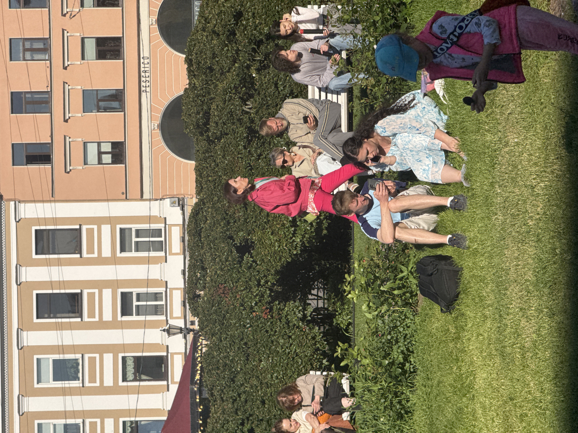 This image captures a gathering of people in a park-like setting, likely in Saint Petersburg, Russia. The sun is shining brightly, casting shadows and indicating a daytime scene. The weather appears to be warm and pleasant.
The subjects are a diverse group of individuals, including men, women, and what appears to be a child. Many are seated or lounging on the grass, while others are on a white bench. The people are dressed casually, suggesting a relaxed social occasion.
Some individuals are engaged in conversation, while others appear to be looking at their phones or resting. One person is holding a microphone, hinting at a possible performance or event. Another is wearing a pink vest over a blue shirt, and a young person in the foreground is wearing a blue cap and a pink vest. A black backpack is visible on the grass.
In the background, a building with a beige facade and white trim, featuring multiple windows, is visible. The building's architectural style, along with the presence of a lush green hedge and trees, provides context for the urban park setting.
There is some visible text in the image: "PESERICO" is written vertically on the building facade. The text "AEROPOSTALE" is visible on the clothing of one of the individuals.