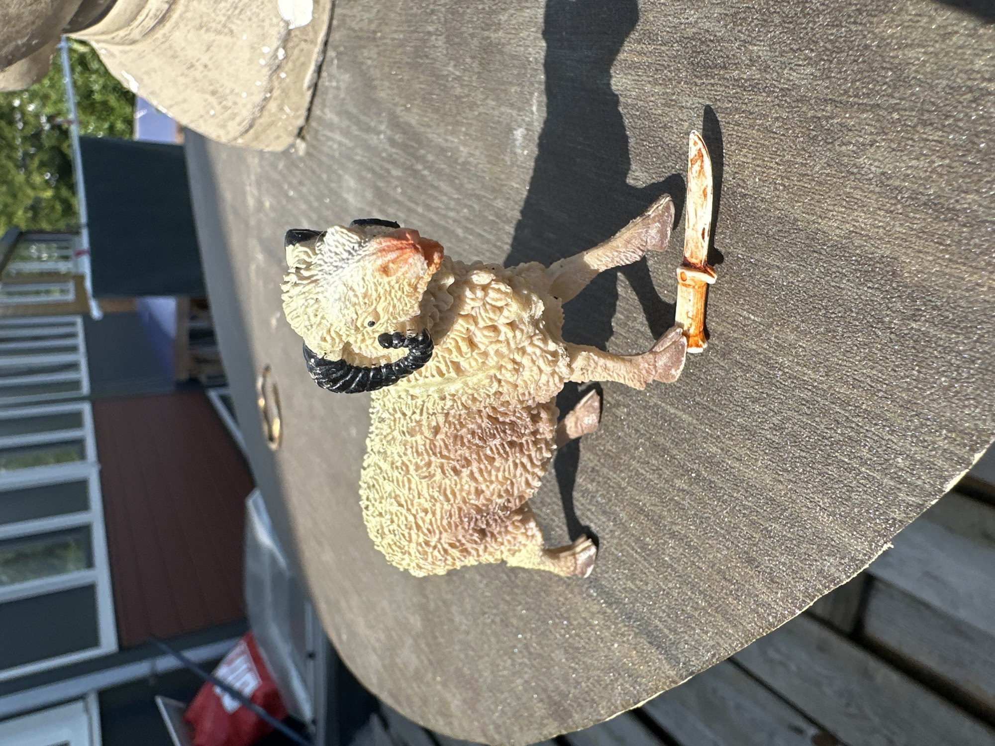 An outdoor, close-up, eye-level shot shows a plastic toy ram standing on a round, textured surface. The ram is light brown and white with black curly horns and appears to be positioned mid-stride, with its front right leg extended. A small, orange and white toy knife lies on the surface in front of the ram's right front hoof. The background is blurred and shows a building with dark red siding and white-framed windows, along with green foliage and a glimpse of a concrete structure to the right. The image appears to have been taken on a sunny day, casting a shadow of the ram to its right. The overall scene is likely a garden or outdoor recreational area in Lelystad, Netherlands.