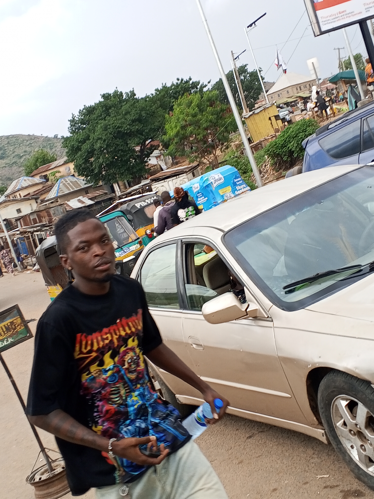 A street scene in Gwong, Nigeria, features a medium-skinned man wearing a black t-shirt with a vibrant graphic print. He holds a water bottle and stands next to a gold-colored sedan car. Behind him are parked tricycle taxis and buildings with corrugated metal roofs. A sign with illegible text is visible near the edge of the image. In the background, other cars and people can be seen on the street, along with a building topped with an antenna. The sky is overcast. A sign displaying a Thursday event, naming "Pastors, Leaders, and Workers" is also displayed.