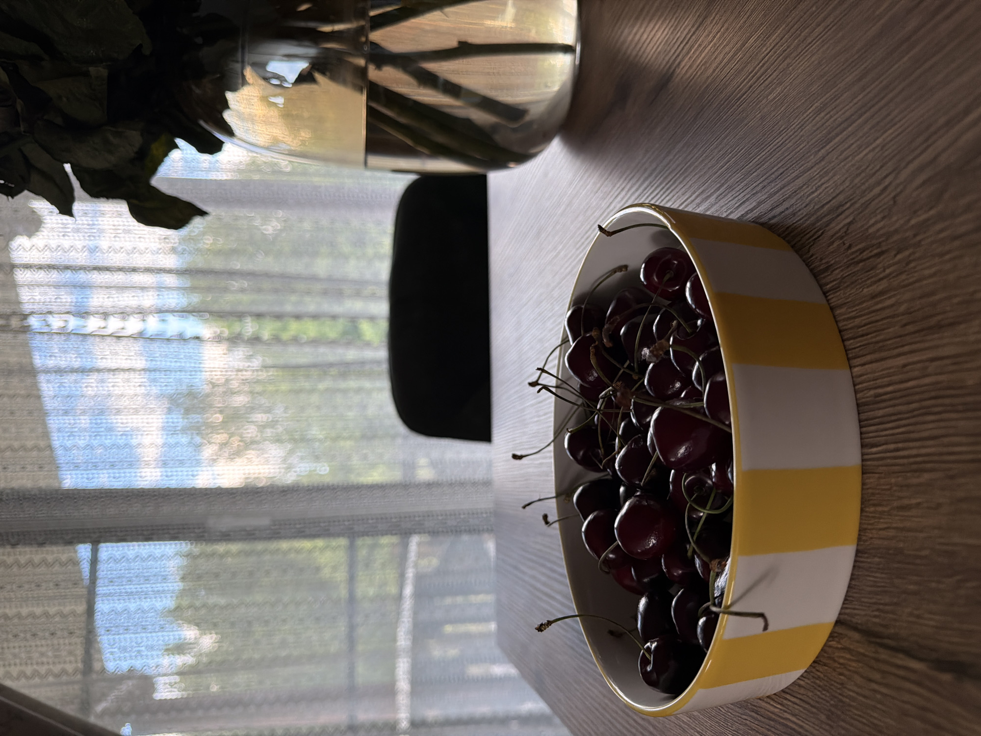 The image displays a close-up of a yellow and white striped bowl filled with ripe cherries. The bowl is placed on a wooden surface, and the lighting suggests it is daytime, with shadows casting diagonally across the wood. To the left of the bowl, a portion of a vase containing what appears to be dried flowers or leaves is visible. In the background, a sheer curtain is drawn over a window, allowing diffused sunlight to filter through, creating a soft, blurry effect of the outdoors, possibly trees and sky. The overall scene is still and domestic, likely an indoor setting in Shashmurka, Russia. There is no visible text in the image.