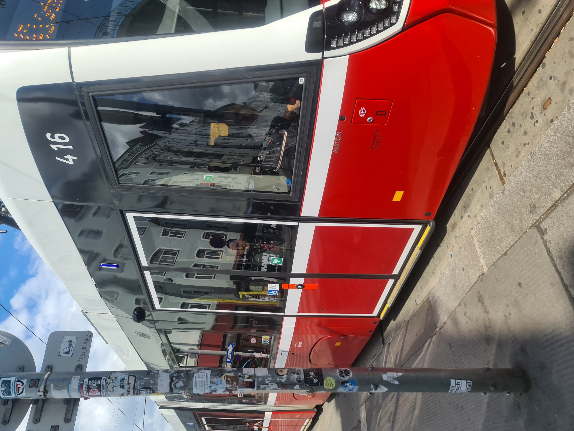 A red and white tram numbered 416 is shown from the side. The tram has large windows that reflect the surrounding buildings and sky. On the side of the tram, the word "ALSTOM" is visible, along with "Wagenmasse 41000 kg" and the letter "D" inside a red square. There are also several stickers on a pole in the foreground, one of which reads "antifascist". The tram appears to be stopped at a tram stop, with tracks visible in the foreground. The overall scene suggests an urban environment in Vienna, Austria, likely on a sunny day given the shadows.