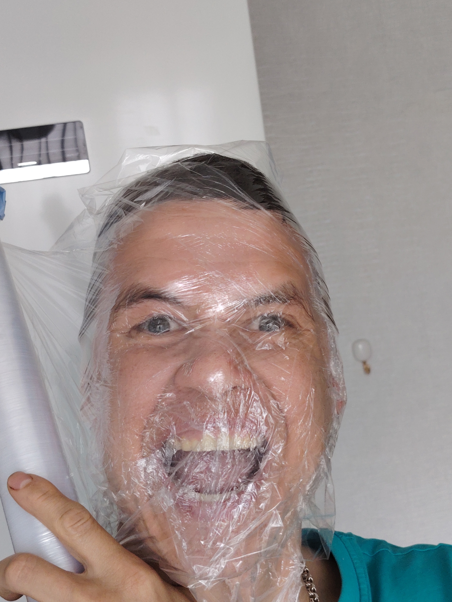 A middle-aged man is shown in a close-up head shot, his face wrapped tightly in clear plastic wrap. His mouth is open in a wide, toothy grin, and his eyes are visible and alert. He is holding a roll of the same plastic wrap in his left hand. He is wearing a teal shirt and a silver chain is visible around his neck. The background is composed of a white wall with a horizontal, dark, rectangular object mounted high on the wall. A small, blurry, round object is also visible on the wall. The image was taken indoors. The city of Chaikovsky, Russia, is not identifiable from this image.