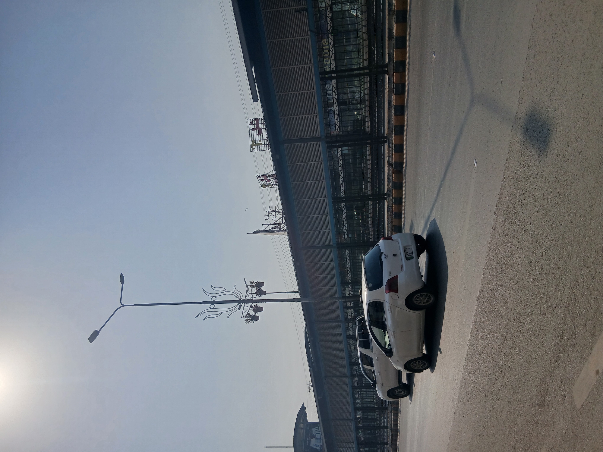 A bright, sunny daytime outdoor scene is depicted in Peshawar City Tehsil, Pakistan. The image focuses on a street with two white vehicles. In the foreground, a white Toyota Vitz is driving on the right side of the road. Behind it, a white pickup truck is also in motion. A large, modern building with a blue facade and louvred windows stretches across the background, with a black metal fence in front of it. A tall street lamp with a decorative element featuring hanging planters stands prominently in the middle ground, to the left of the vehicles. The sky is clear and bright with the sun at the top left, casting shadows on the asphalt road. The overall impression is one of an urban environment on a clear day.