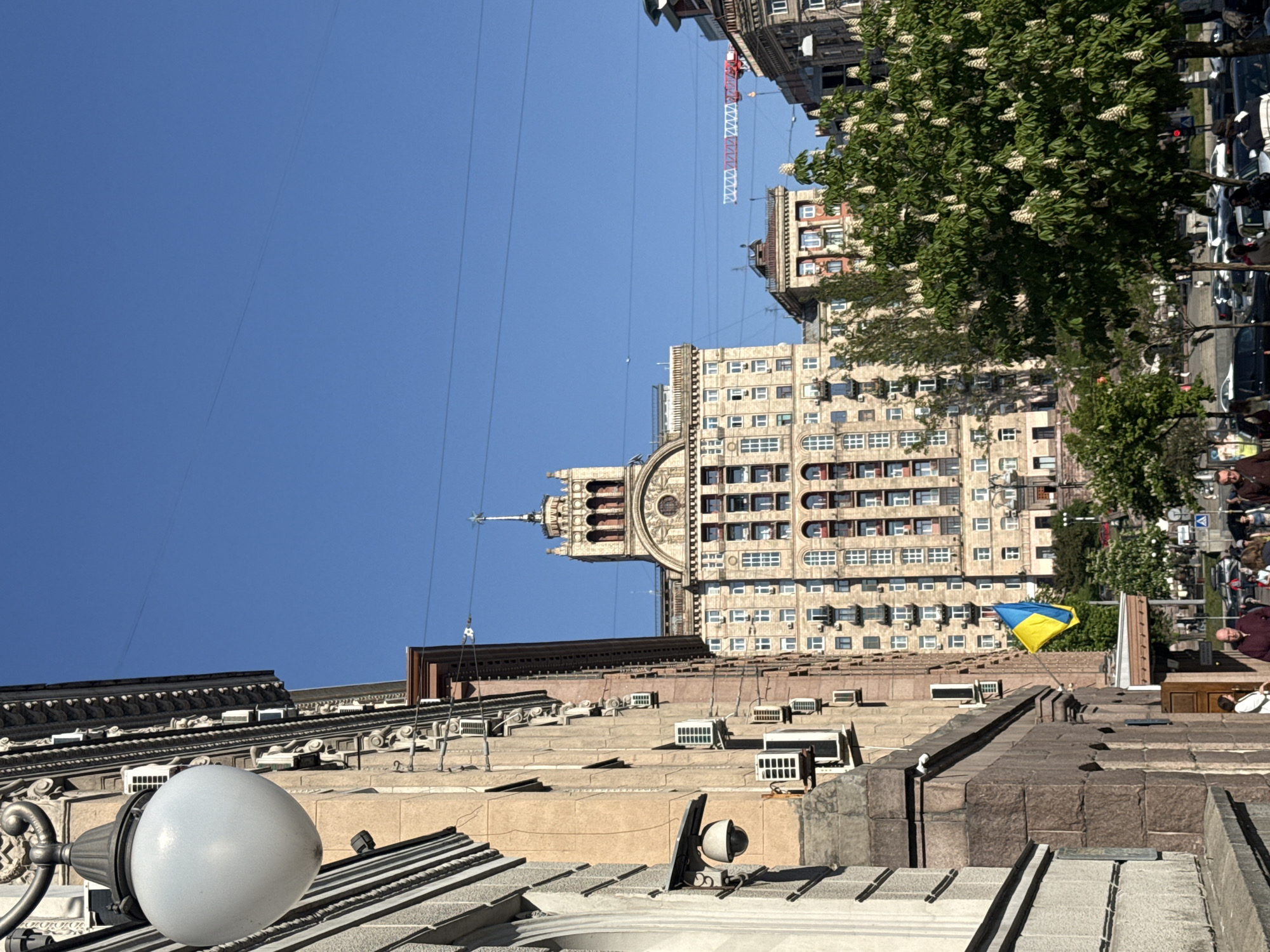The image, taken from a low angle, shows the cityscape of Kyiv, Ukraine. The sky is clear and blue. In the foreground, there is a building with a visible roof. Various air conditioning units are mounted on the building's facade. A Ukrainian flag is visible on the roof. The background features a large, multi-story building with architectural details including a clock tower and rows of windows. A tree with white flowers is partially visible on the right side of the image, along with a line of cars and some people. The electrical cables are visible against the blue sky.