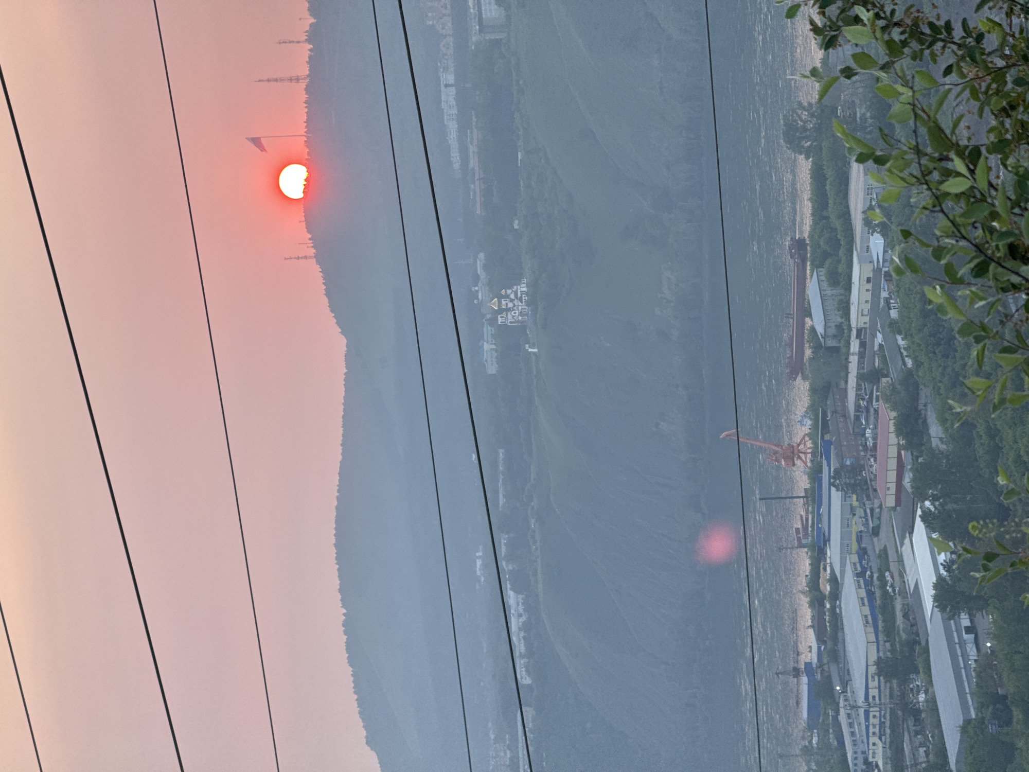 This media file depicts a scenic view of a river valley with a setting sun. The sun, a vibrant orange, is partially obscured by a mountain range on the left side of the frame, casting a warm glow across the sky and creating a soft, hazy atmosphere. The sun appears to be setting, as indicated by its low position and the rosy hue of the sky.
Dominating the foreground are several dark, taut power lines, stretching diagonally across the image. These lines create a strong visual element, framing the landscape and suggesting a human presence or infrastructure within the natural setting.
In the middle ground, the river is visible, its surface shimmering with the reflected light of the setting sun. Along the riverbanks, a cluster of industrial buildings and structures can be seen. A large red crane stands prominently, hinting at port activities or construction. The architecture of the buildings suggests a commercial or industrial area, with various warehouses and factory-like structures.
Further in the distance, the slopes of the mountains are covered in dense vegetation, appearing as a dark green mass against the lighter sky. The overall impression is one of a vast, somewhat rugged landscape with signs of industrial development along the river. The image conveys a sense of tranquility and natural beauty, juxtaposed with the utilitarian presence of human activity. The time of day is clearly late afternoon or early evening, given the sun's position and the soft lighting. The weather appears clear, with the haze likely due to atmospheric conditions or the distance. The location is implied to be a river valley with surrounding mountains, consistent with the description of Krasnoyarsk, Russia. There is no visible text in the image.