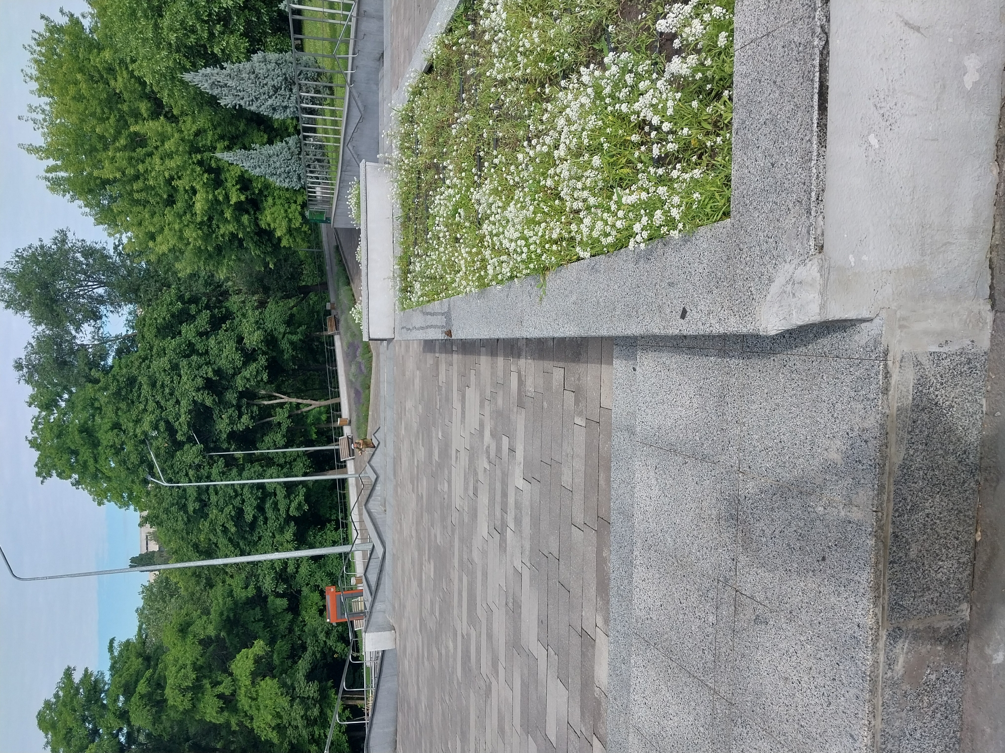 The image shows a stone staircase and walkway in a park-like setting with trees and flowerbeds. A person in military uniform is visible walking up the stairs. A small, orange structure that appears to be a kiosk or information booth is located in the background to the left. There are also two benches and two lampposts in the scene. The environment appears to be outdoors, likely a public park or plaza. The weather appears to be clear and sunny, with some clouds in the sky. The overall impression is one of a well-maintained outdoor public space.