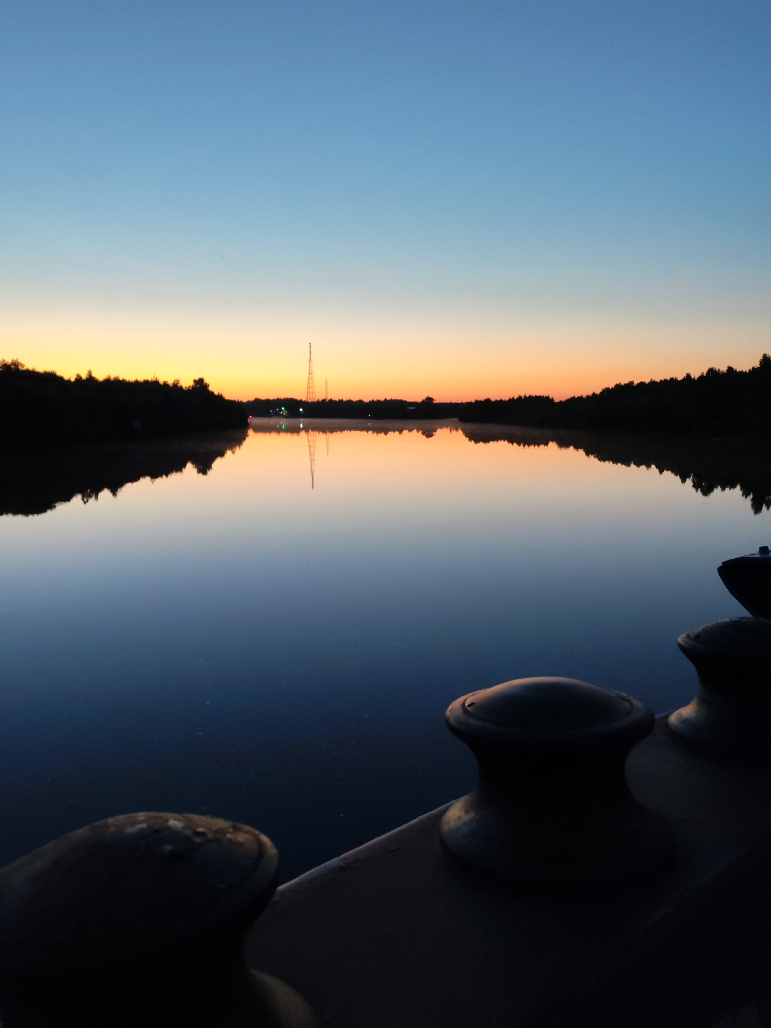 This image captures a serene twilight scene over a calm body of water, likely a river or lake, at Annensky Most, Russia. The sky transitions from a deep blue at the top to a vibrant orange and yellow gradient near the horizon, indicating either sunrise or sunset. A silhouetted treeline borders the far bank of the water, with a tall radio tower visible against the colorful sky. The water itself is a mirror, perfectly reflecting the sky and the distant treeline, creating a symmetrical and peaceful composition. In the foreground, the dark silhouettes of two mooring bollards, characteristic of a harbor or dock, are visible on the edge of what appears to be a boat or pier. The overall atmosphere is tranquil and still, with no people or animals present. The lighting suggests low light conditions, typical of dawn or dusk.
