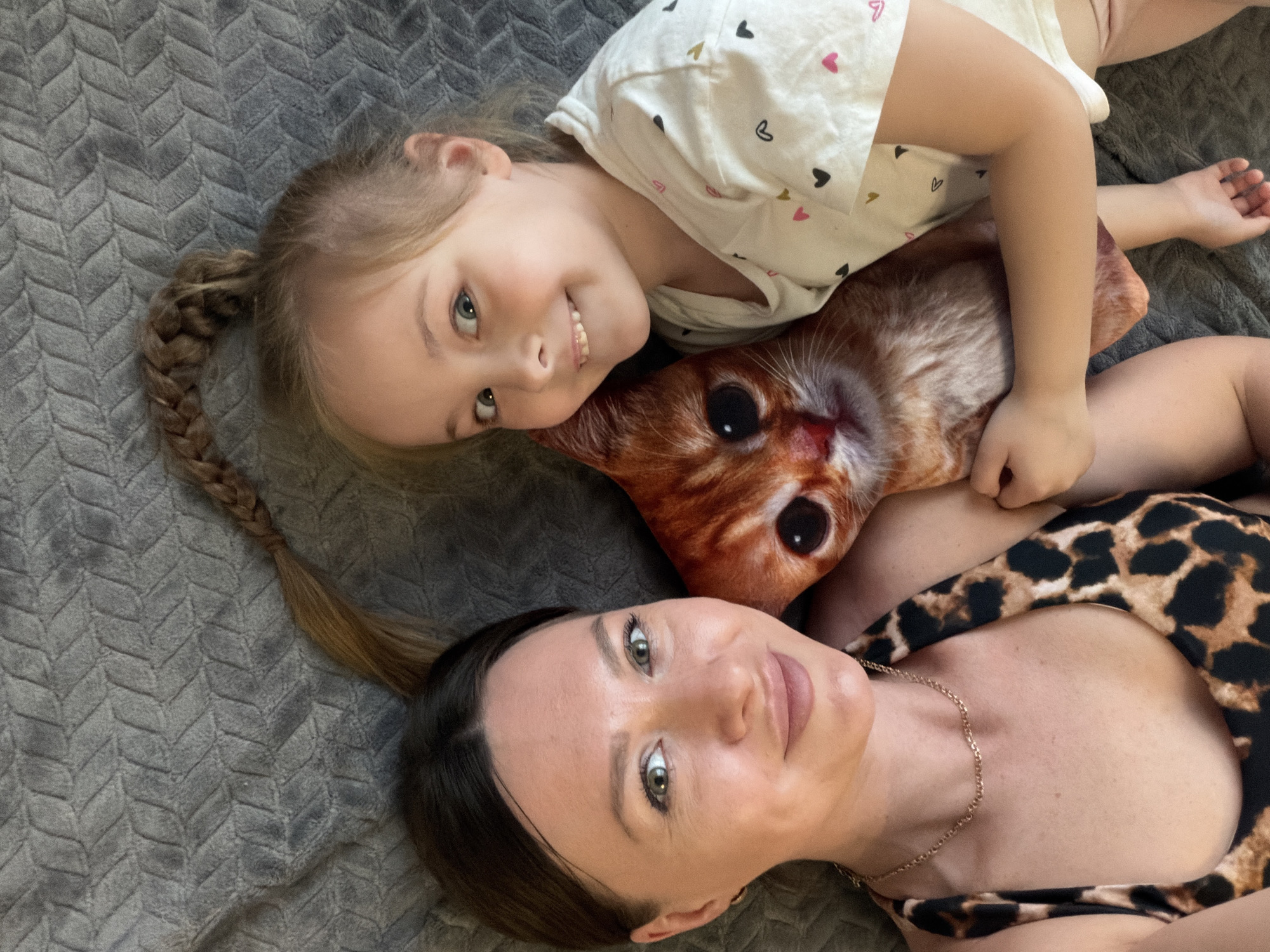 A young child and a woman are lying on a gray textured blanket or bedspread. The child, positioned at the top, has blonde hair in a braid and is smiling at the camera. She wears a white short-sleeved shirt with heart patterns. Beneath her, between her and the woman, is a pillow shaped like a cat. The woman, positioned at the bottom, also faces the camera. She has dark hair pulled back and is wearing a leopard print top with a gold necklace. The environment appears to be an indoor setting. There is no information to place this image in Rostov-on-Don, Russia.