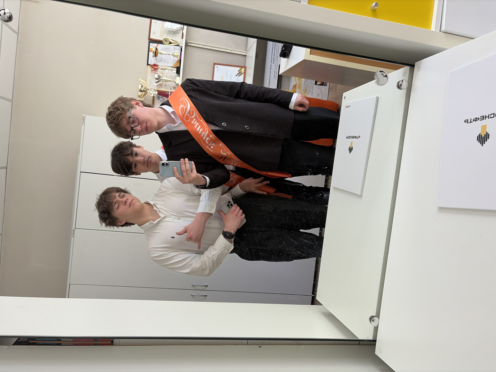 Three young men are reflected in a mirror. The man at the top wears glasses, a dark blazer, and an orange sash printed with Cyrillic text. The middle man holds a cell phone to take a picture. The third man wears a white dress shirt and dark pants and is pointing down. The reflection shows they are in a room with white cabinets and a yellow drawer visible above. Behind them are trophies and documents on the wall. A sign in Cyrillic is fixed on the white panel. The logo on the sign indicates this is BOCHFELP, which locates the scene in the city of Krasnoyarsk, Russia.