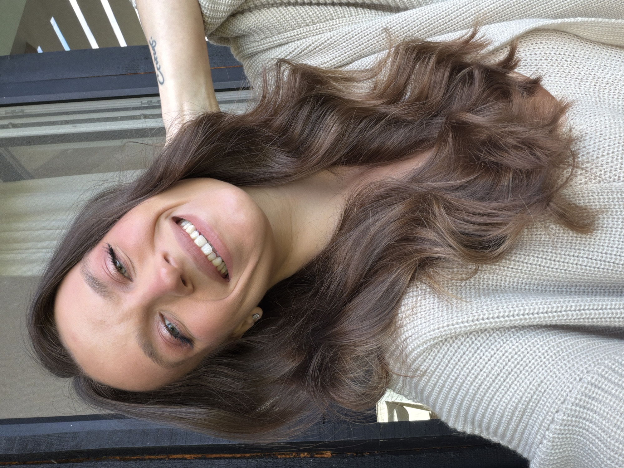 A woman with long brown hair is smiling at the camera. She is wearing a beige knit sweater. The background includes a dark wooden surface and some light coming through slats. No information suggests the location of Prudki, Russia.
