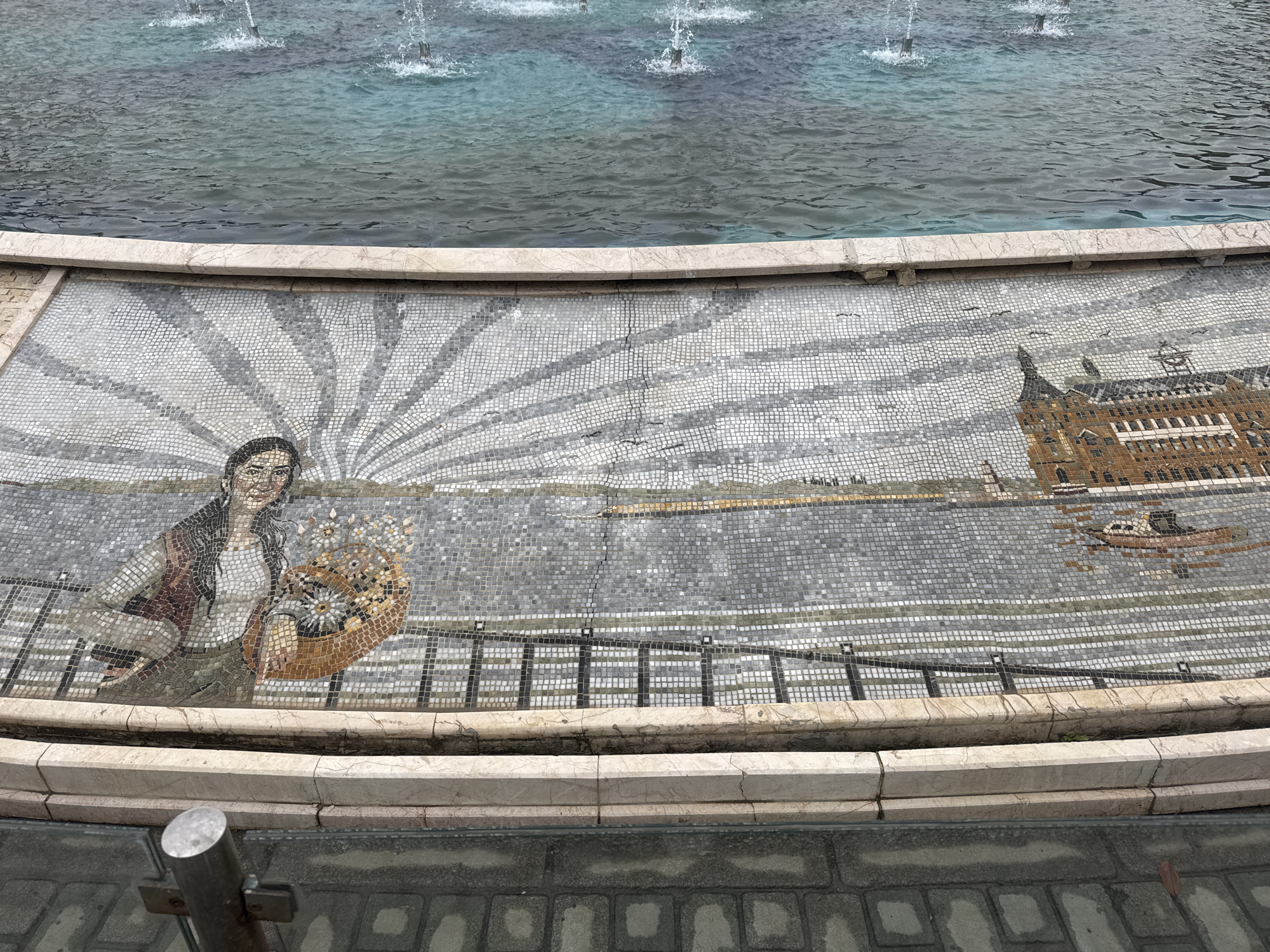 The image shows a mosaic on the side of a fountain in Istanbul, Turkey. The mosaic depicts a woman holding a basket of flowers, a body of water, and the Haydarpaşa Train Station. The fountain has multiple jets of water spraying upwards. The mosaic is made of small, square tiles. The surrounding area is paved with gray bricks. A metal pole is visible on the left side.