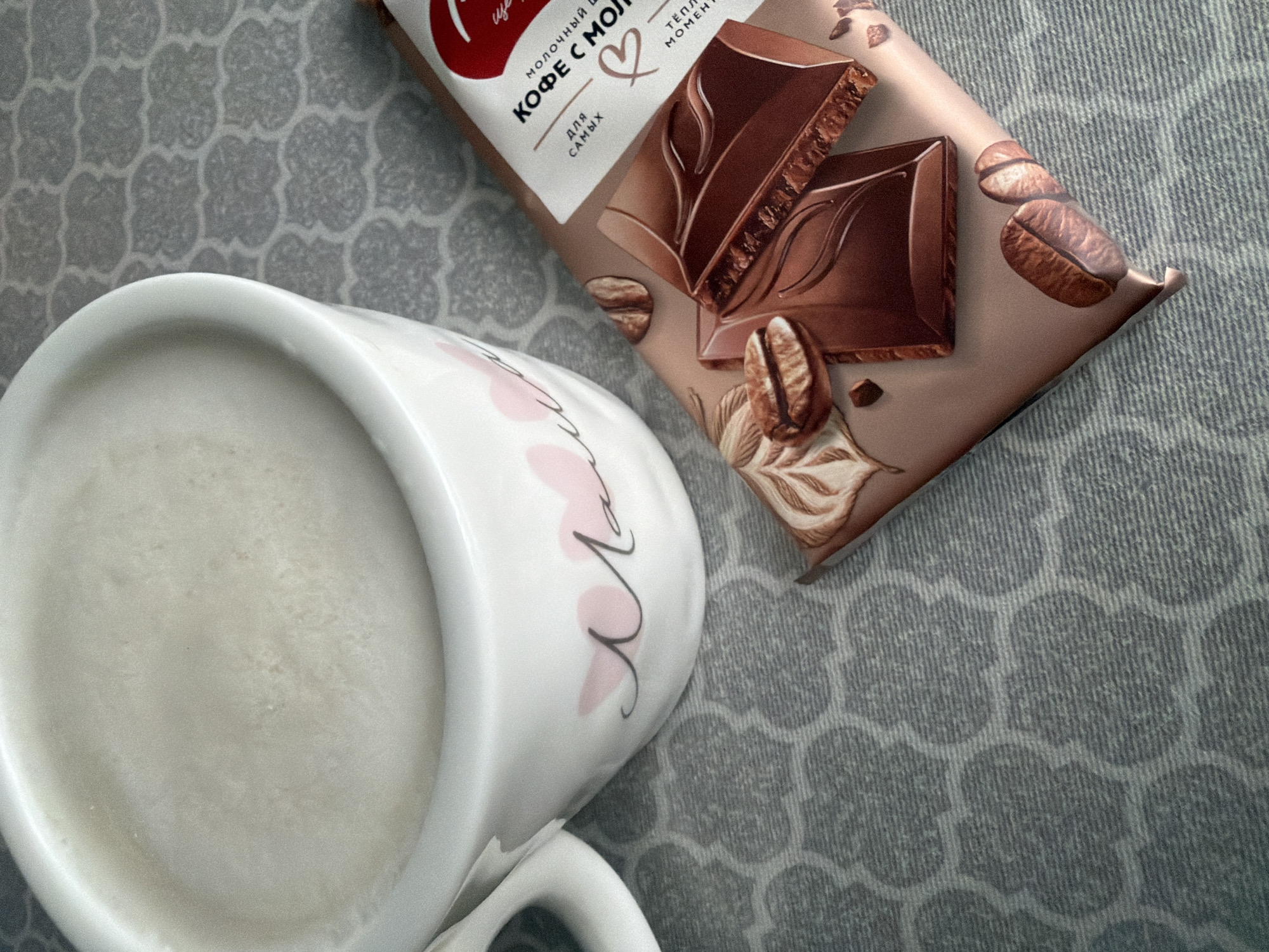 The image depicts a mug of a white liquid, likely milk or a milk-based beverage, and a chocolate bar with coffee flavor against a patterned surface. The chocolate bar is positioned diagonally in the upper right of the frame. The mug, featuring pink floral decorations and possibly Cyrillic script, is positioned in the lower left. The patterned surface is light grey. No people are visible. There is no indication of the location being Votkinsk, Russia, in the image itself.