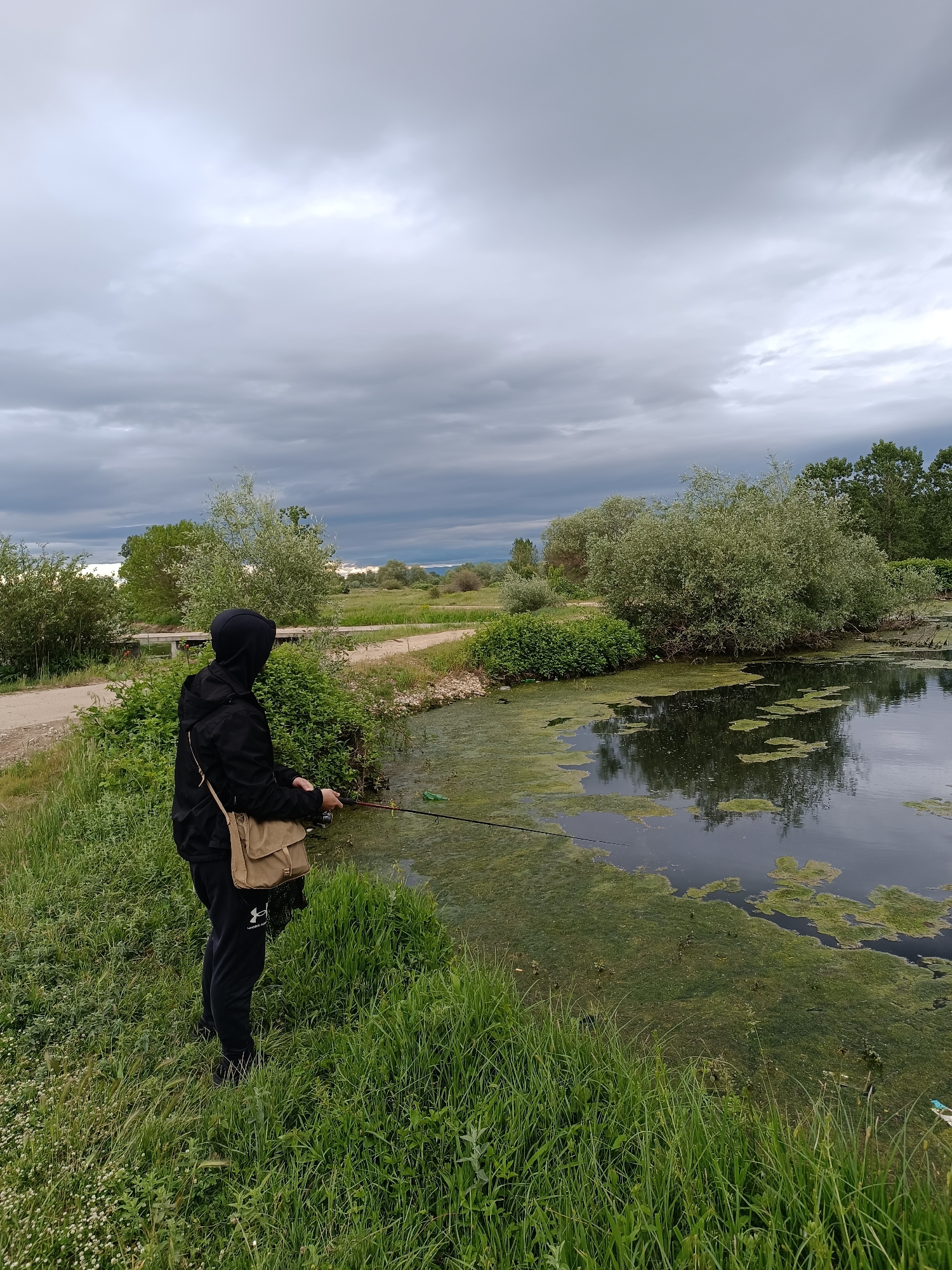 A person wearing a black hooded jacket and black pants with an Under Armour logo is fishing in a body of water. The person has a tan-colored messenger bag slung over their shoulder and is holding a fishing rod. The water is covered with green algae or vegetation. The surrounding area includes grassy banks, bushes, and trees. In the background, there is a path, more vegetation, and a cloudy sky. The scene appears to be outdoors, potentially in a park or natural area. The location is Zlokuchene, Bulgaria.