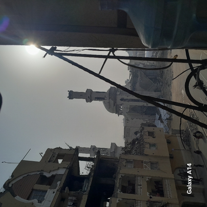 Rubble-strewn buildings stand in disrepair, possibly the result of conflict or disaster. A mosque's minaret rises amidst the devastation. The scene is captured from an elevated vantage point, with foreground objects partially obscuring the view. The sky is hazy or overcast, contributing to the somber atmosphere. The general impression is one of destruction and desolation. A water bottle can be seen in the upper right corner. The camera used to take the picture is identified as a Galaxy A14 in the lower right corner.