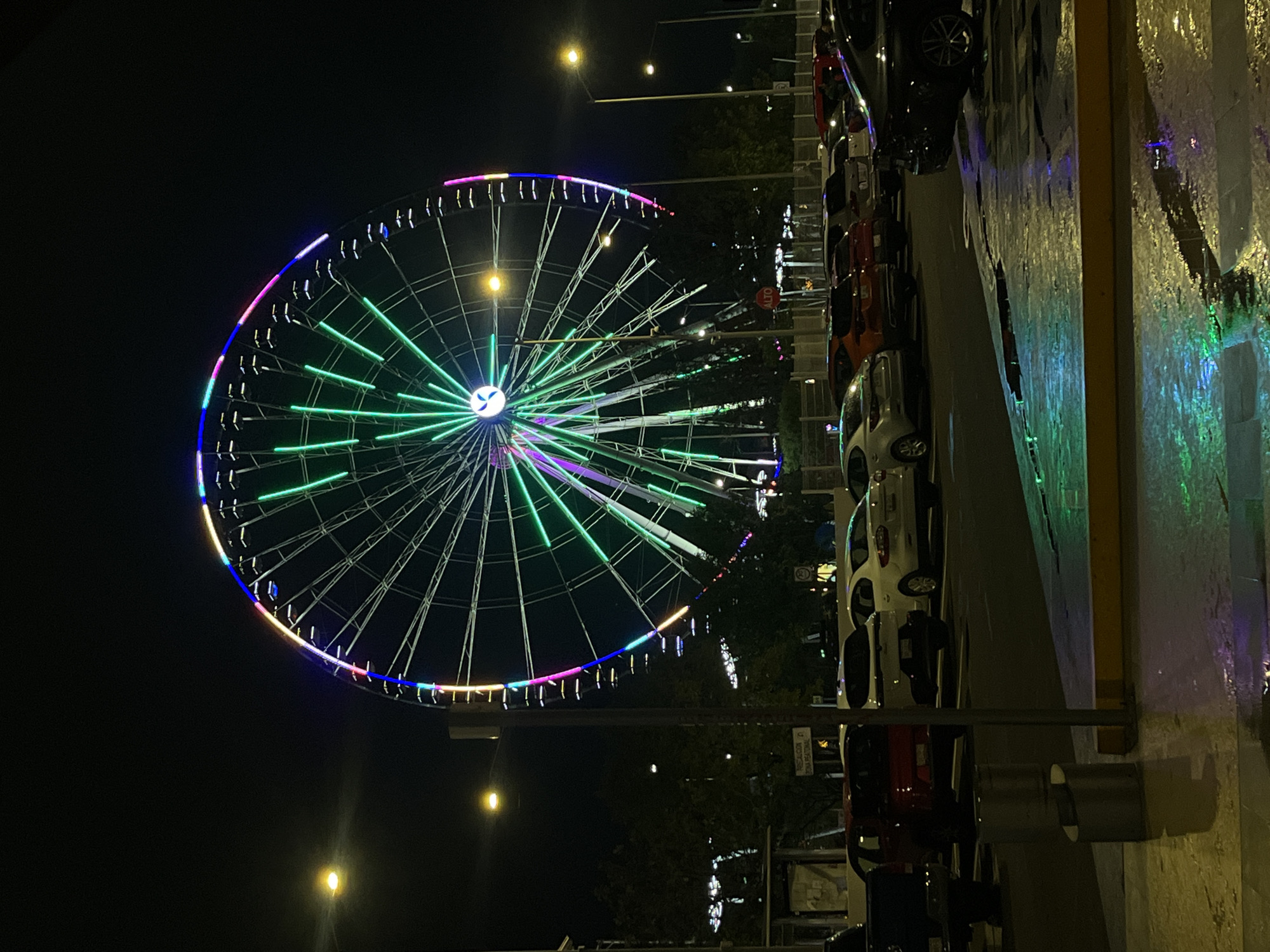 The image depicts a Ferris wheel at night, brightly lit with green spokes and a multicolored rim. Cars are parked along the right side of the frame, and streetlights illuminate the scene. The location appears to be urban, with a wet pavement reflecting the lights. The absence of people and distinct landmarks makes it difficult to identify the specific city.