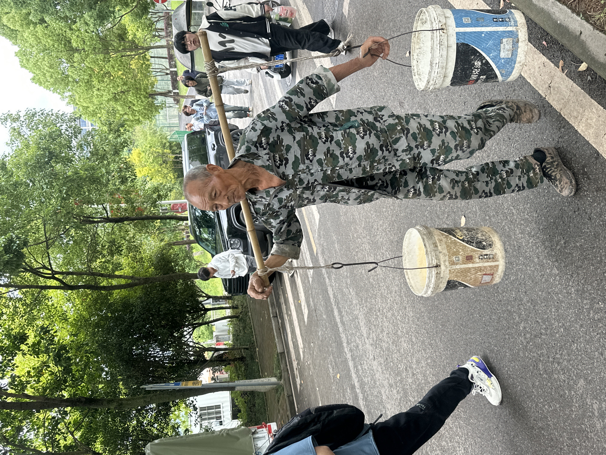 This image, taken in Jiangjin, China, depicts an outdoor street scene during what appears to be an overcast daytime. The perspective is rotated, presenting the subjects vertically.
The central figure is an older man, dressed in a full camouflage uniform, including a jacket, pants, and shoes. He is carrying two white, cylindrical buckets suspended from a wooden or bamboo pole across his shoulders, a traditional method of transporting goods. His head is slightly bowed, suggesting focus on balancing the load. The buckets appear to be used industrial containers; one has distinct blue and white markings with the Chinese characters "固安夜" (Gù'ān yè) visible, while the other shows tan/brown marks.
In the background, several other people are present. A young man in a black and white jacket stands near a scooter, observing the scene. Other individuals are further back, some near a black SUV. A partial view of a person's leg and foot in dark pants and white sneakers is visible in the foreground.
The setting includes a paved road with white markings, lush green trees lining the roadside, and faint architectural structures in the distance. The overall impression is a candid moment captured on a street, highlighting a traditional method of carrying goods amidst a modern environment.