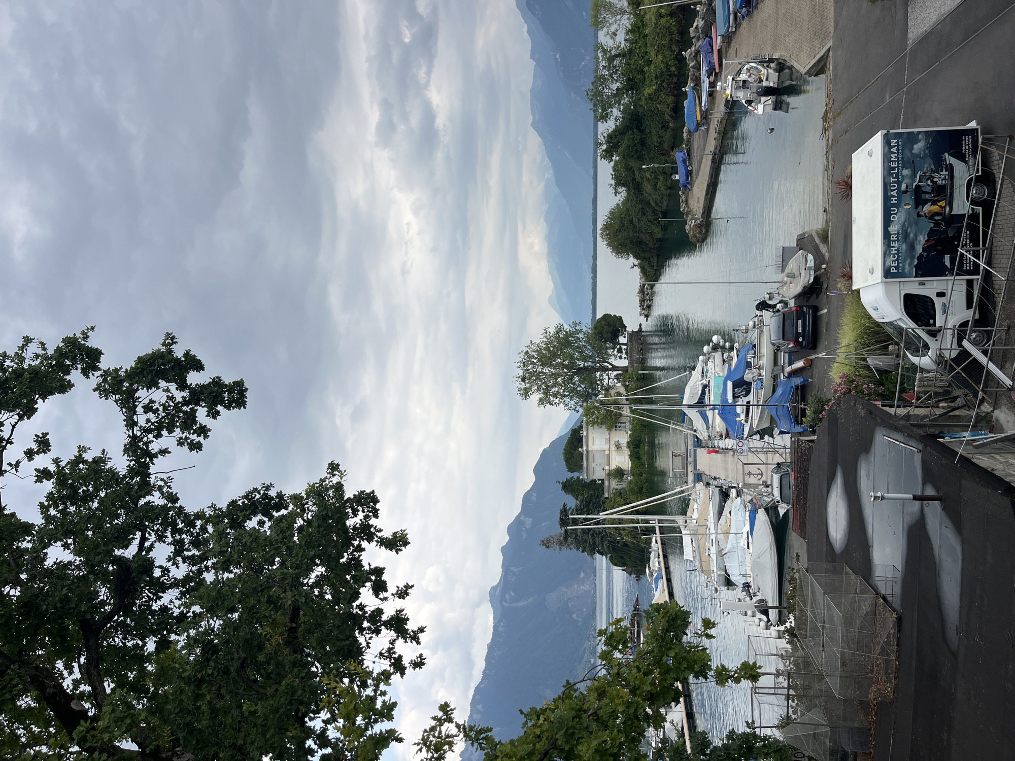 The image depicts a serene view of the port of Montreux, Switzerland, on a cloudy day. In the foreground, a dark rooftop with some puddles is visible, alongside several wire cages. A white van with the advertisement "PÉCHERIE DU HAUT-LÉMAN ARTISANS PÊCHEURS" is parked on the right. Beyond the rooftop, a harbor filled with various sailboats and smaller motorboats lines the water. Some boats are covered, while others are partially visible. The calm water of Lake Geneva stretches out, with a small island featuring a white building and a tree situated in the middle of the lake. Majestic mountains form the backdrop, their peaks softened by the overcast sky. The overall scene is tranquil, showcasing the natural beauty and the recreational activities associated with the port.