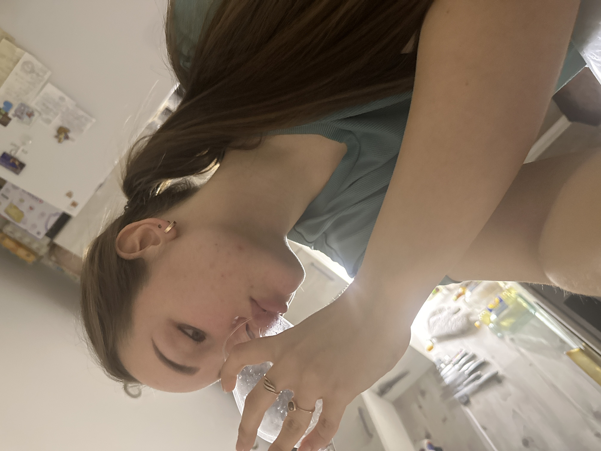 A young woman with long brown hair is tilted toward the camera, drinking from a clear glass. She wears a light blue shirt and gold rings on her fingers. The background includes a refrigerator covered with papers, a countertop with various items, and a white sink. The location cannot be determined from the image and there is no evidence the image was taken in Chaikovsky, Russia.