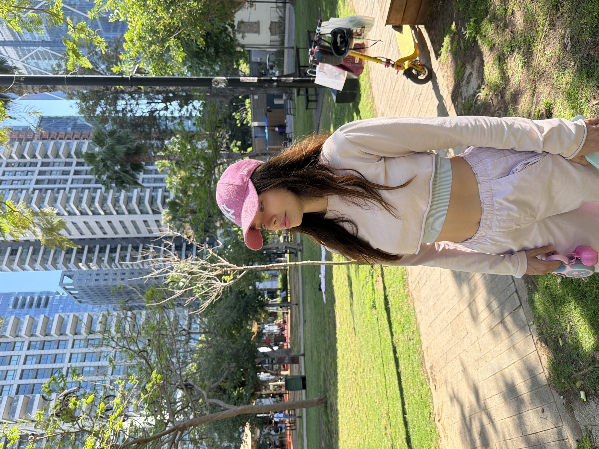A young woman in a pink baseball cap, pale pink long-sleeved shirt, and pale pink sweatpants stands on a brick path within a park. The path is adjacent to green grass. Behind her, a child's yellow scooter rests near a park bench. High-rise buildings can be seen in the distance, indicating an urban setting. The location is likely in the city of Tel-Aviv, Israel.