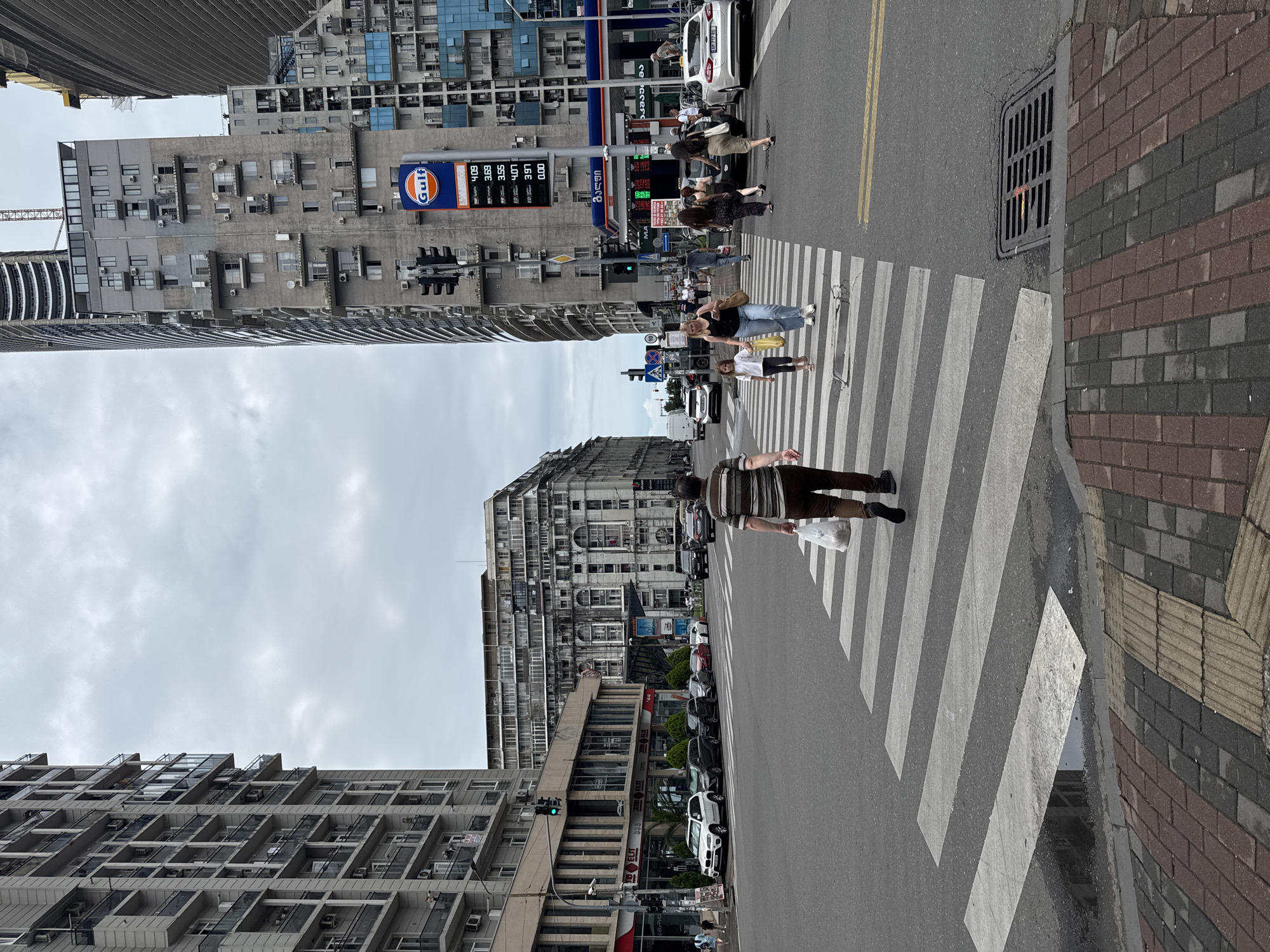 The image depicts a street scene in Batumi, Georgia, on a cloudy day. The foreground features a pedestrian crossing with several people walking on it. A man in a striped shirt and brown pants is walking away from the camera, carrying a white plastic bag. To his left, a woman in a black tank top and jeans walks with a young girl in a pink shirt, both holding yellow bags. Other pedestrians are visible crossing the street or walking on the sidewalks.
The street is lined with modern and older buildings. On the left, a tall, contemporary residential building with balconies and air conditioning units dominates the view. Across the street, a large, older building with an ornate facade and numerous windows is visible. To the right, another modern building with a Gulf gas station sign and a digital display showing fuel prices stands prominently. The gas station has a blue canopy and pumps. Cars are parked along the street and some are driving through the intersection. The road surface shows a zebra crossing, road markings, and a storm drain. The overall environment appears to be a busy urban intersection in Batumi.