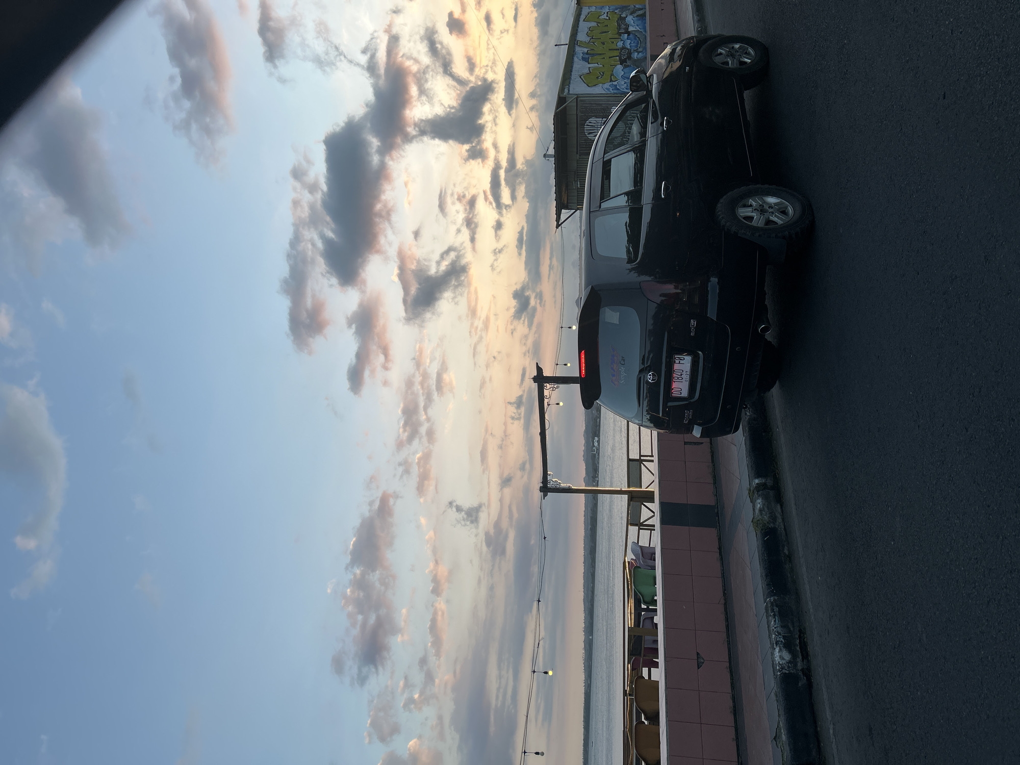 This is a medium shot, eye-level, vertical photograph taken from a road at dusk in Bantaeng, Indonesia. The scene depicts a black Toyota Innova car parked on the right side of a paved road. In the background, there is a body of water, likely the sea, with a distant shoreline visible. Along the waterfront, there is a row of outdoor seating with tables and chairs, suggesting a restaurant or cafe. Overhead, string lights are suspended between poles. The sky is filled with scattered clouds, tinged with the colors of sunset. A small, simple structure with graffiti is visible to the right of the car. The overall impression is a serene, coastal evening scene.