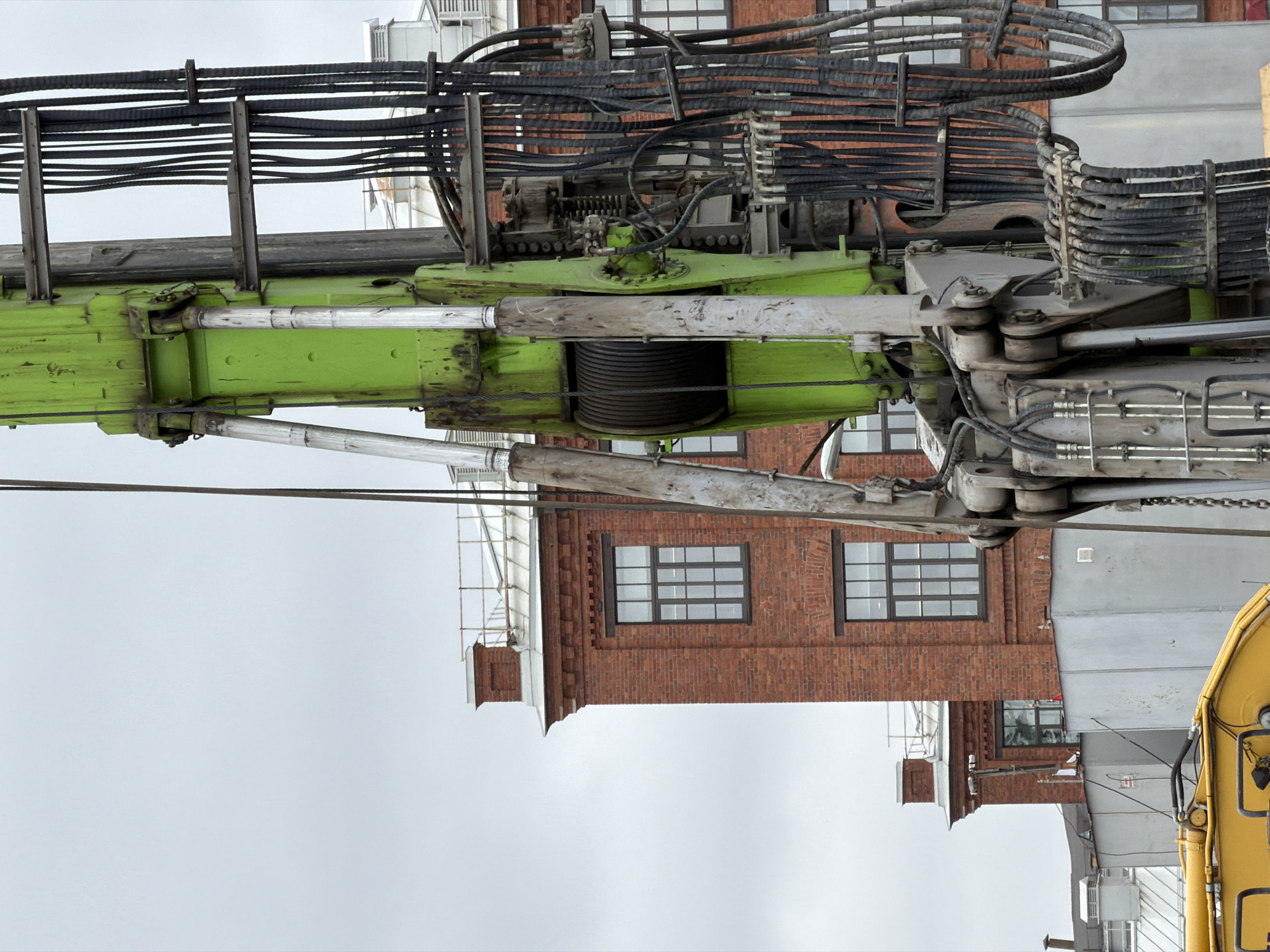 The image shows a close-up of a large green construction crane with visible hydraulic lines and cables. In the background, there is a red brick industrial building with multiple windows. The sky is overcast. The scene is likely a construction site in Saint Petersburg, Russia, given the architectural style of the building. No people are visible in the image.
