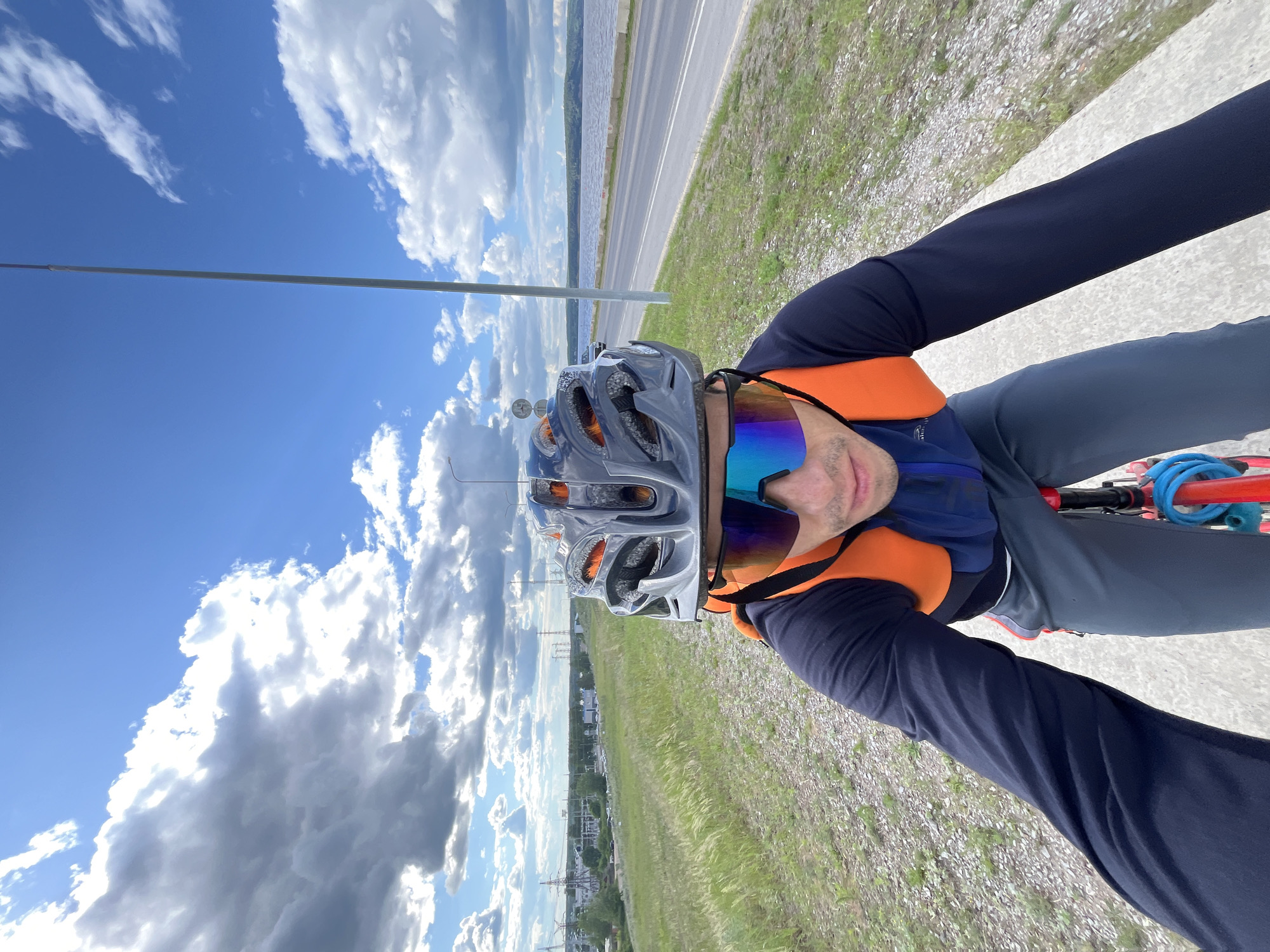 A male cyclist wearing a grey helmet, bright orange vest, dark blue long-sleeved shirt, and grey pants rides a bicycle on a paved path. The cyclist is looking at the camera and is wearing reflective sunglasses. In the background, there is a road, a body of water, and a cloudy blue sky. The scene appears to be on the outskirts of Chaikovsky, Russia, near the Kama River, a common location for recreational cycling and outdoor activities. The presence of industrial infrastructure in the distance, such as power lines, suggests the proximity of an urban or industrial area.