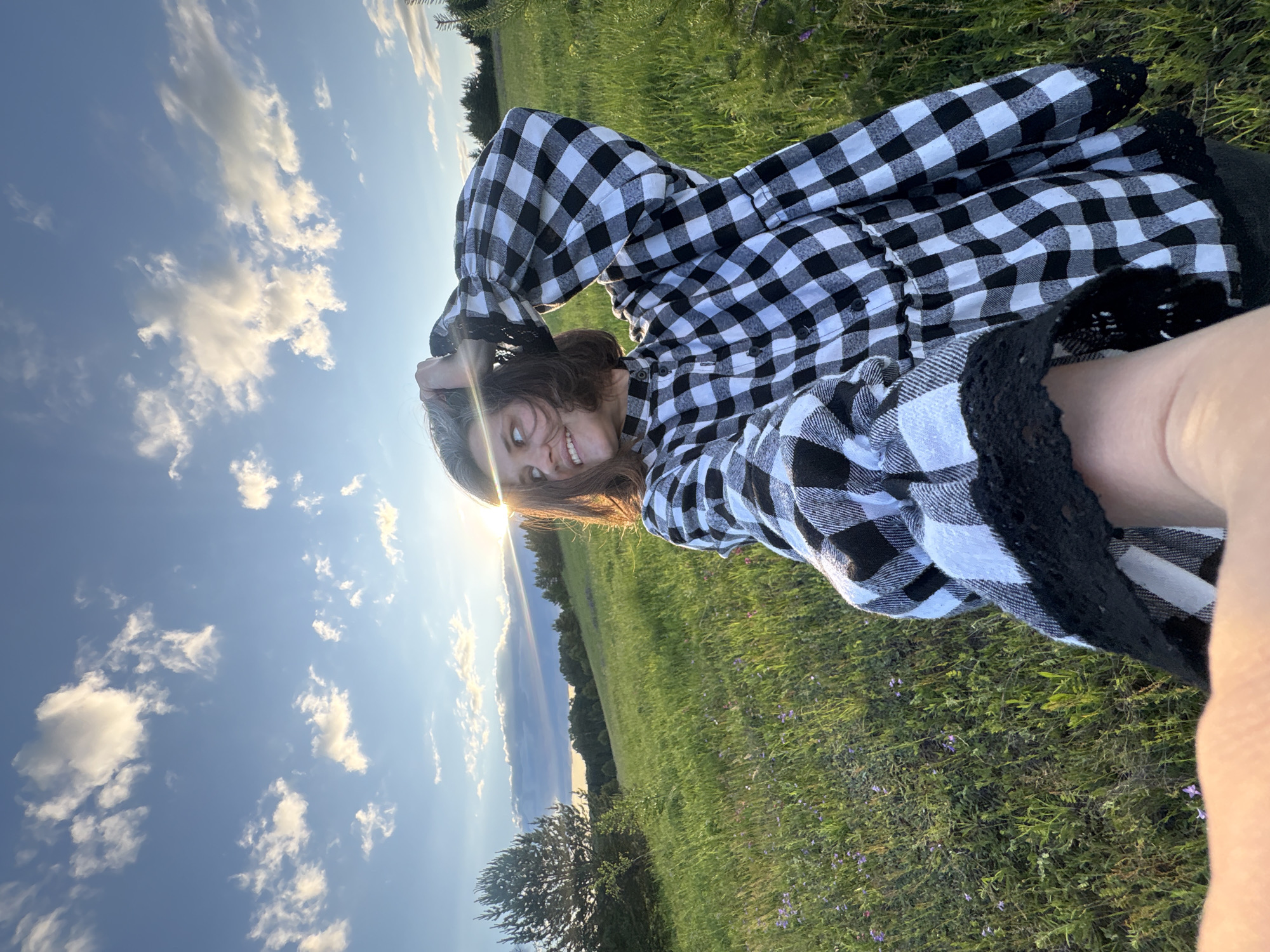 A medium shot captures a young woman with short brown hair standing in a grassy field at sunset. She is wearing a black and white checkered dress with puffed sleeves and black lace trim. The woman is holding her left arm up, with her hand in her hair, and looking to her left with a smile. The sun is low in the sky, casting a bright glare and long shadows across the field. The background consists of rolling hills, trees, and a blue sky with scattered clouds. The location is an outdoor setting in an unspecified rural area.