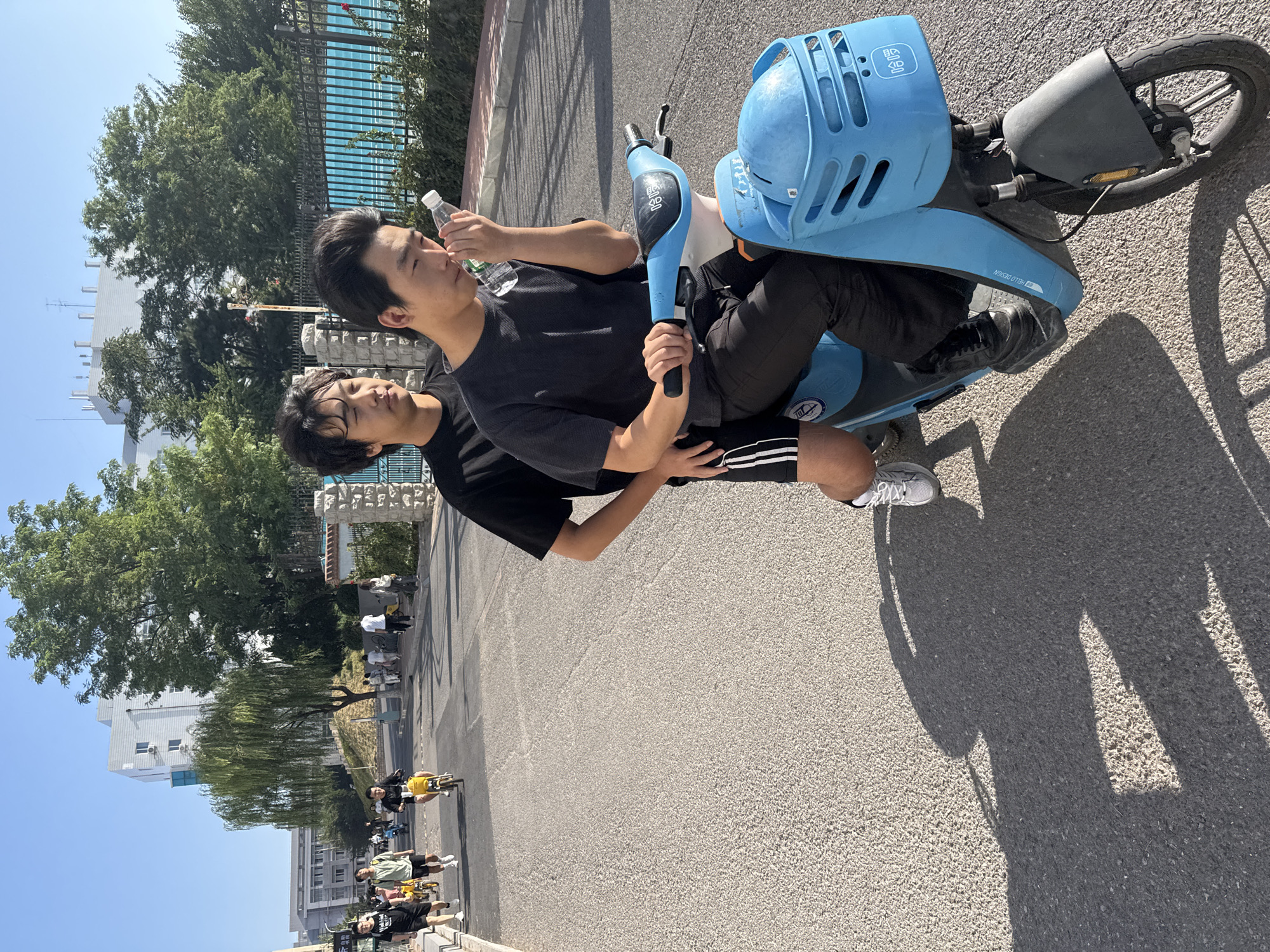 Two young Asian men are riding a bright blue shared electric scooter on a paved road in Zhongtun, China. The man in front is driving, holding the handlebars with his right hand and a plastic bottle of water with his left, bringing it to his face. He wears a dark gray t-shirt and black pants with white stripes on the sides of the legs, and black sneakers. The man behind him, dressed in a black t-shirt and black shorts with white stripes, has his arms around the driver. Both have their heads slightly bowed. The scooter has a basket at the front with some text in Chinese characters. The road is asphalt, with a white line marking the edge. In the background, other people are visible riding yellow electric scooters, walking, and standing on the sidewalk. There are trees and buildings, including a modern white building with blue windows. The sky is clear and blue, indicating it is a sunny day. The shadows of the scooter and the people are cast on the road.