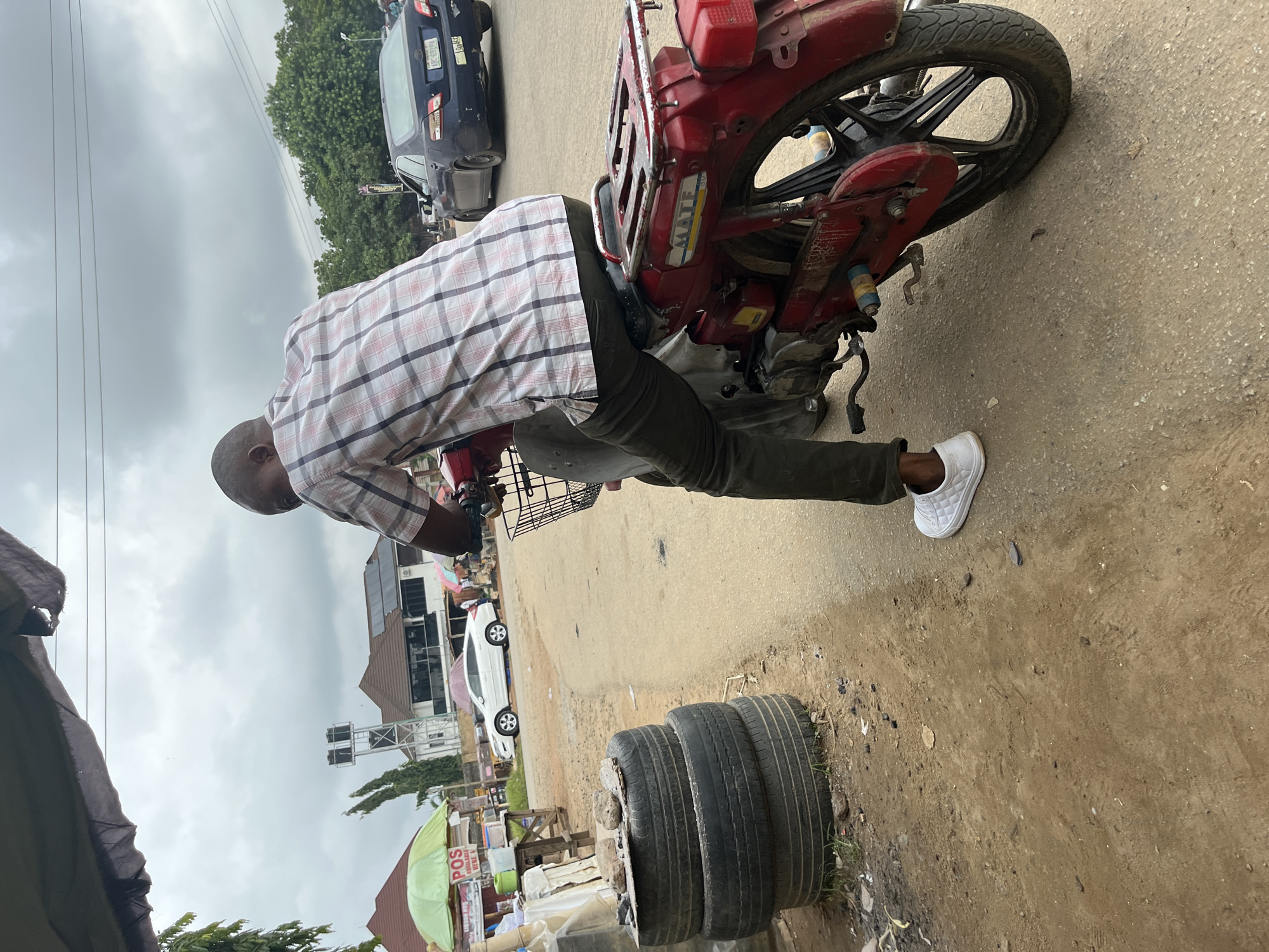 A person wearing a checkered shirt and dark pants sits on a red motorcycle. The motorcycle has a black basket on the front and a metal rack on the back. The person is wearing white sneakers. In the background, there are buildings, trees, and cars on a street under a cloudy sky. A stack of old tires is visible on the left side of the image. The scene appears to be in a street or market area.