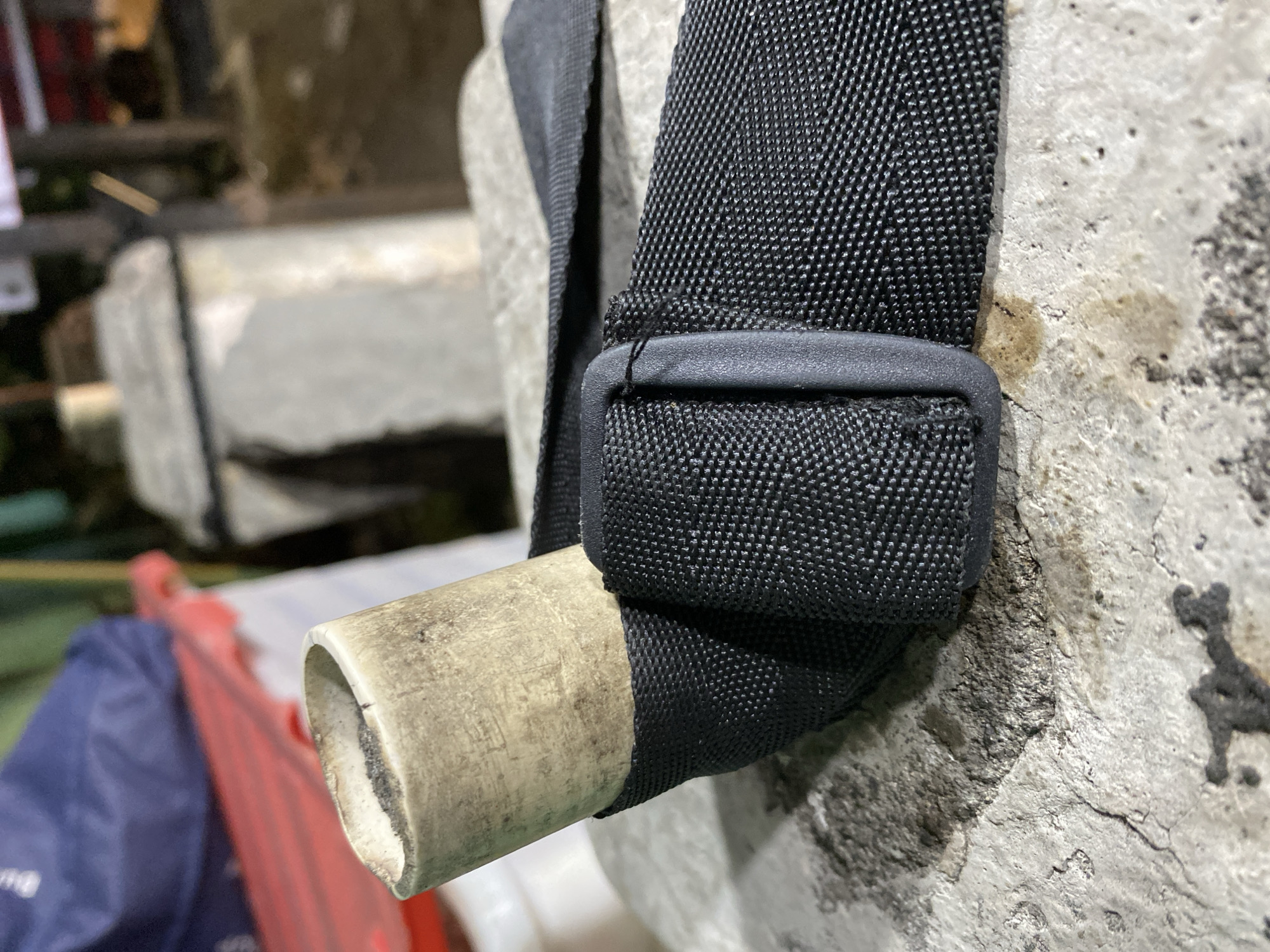 A close-up, low-angle shot focuses on a cream-colored, hollow pipe with a rough, unfinished end, positioned vertically at a slight tilt. A black, textured fabric strap wraps around the base of the pipe, secured by a black plastic buckle. The strap appears to be made of durable material, possibly nylon or polyester, with a visible grid pattern. The pipe and strap are resting on a textured, light-gray surface that resembles concrete or stone, with dark smudges and rough patches. In the background, out of focus, are elements of an outdoor environment, including what appears to be a blue and red tarp or bag, and a light-colored pillar or structure. The lighting suggests it is either dawn or dusk, or an indoor area with artificial lighting. The scene is devoid of human presence or activity, presenting a static arrangement of objects. The location is described as Kasihan, Indonesia.