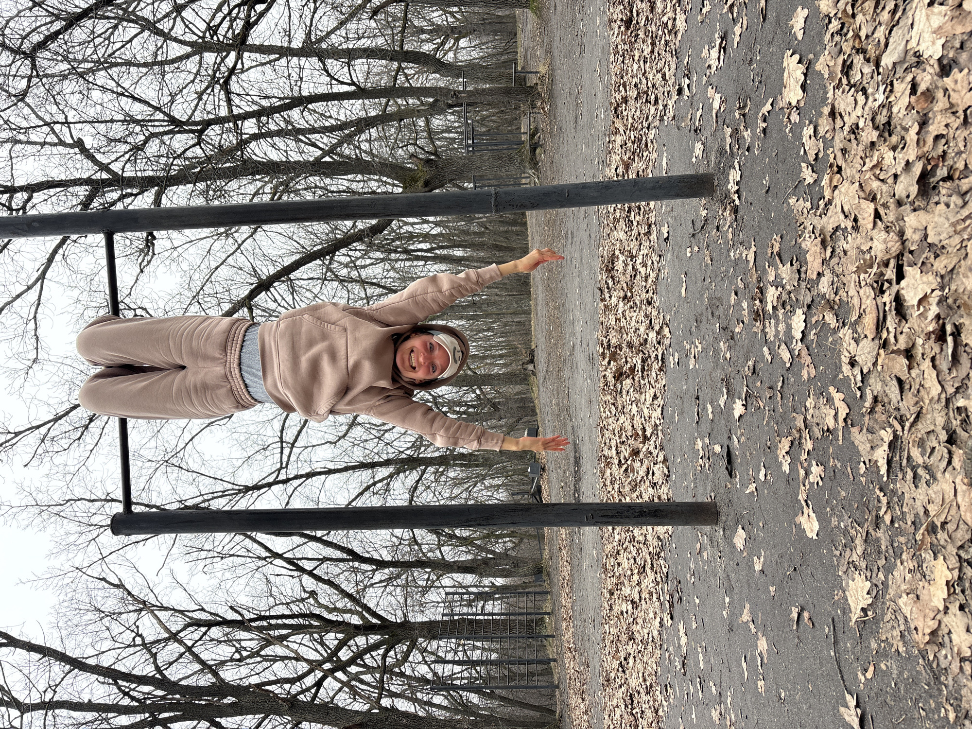 A woman hangs upside down on a horizontal metal bar in a park in Ulyanovsk, Russia. She is wearing a light brown hooded sweatshirt and matching sweatpants with a gray undershirt visible. Her arms are extended to the sides, and she is smiling. The metal bar is supported by two vertical posts. Behind the bar are bare tree branches, indicating it is likely winter or early spring. To the right, the ground is covered in fallen leaves, and a path extends into the distance. Another set of parallel bars can be seen further along the path, suggesting a workout area.