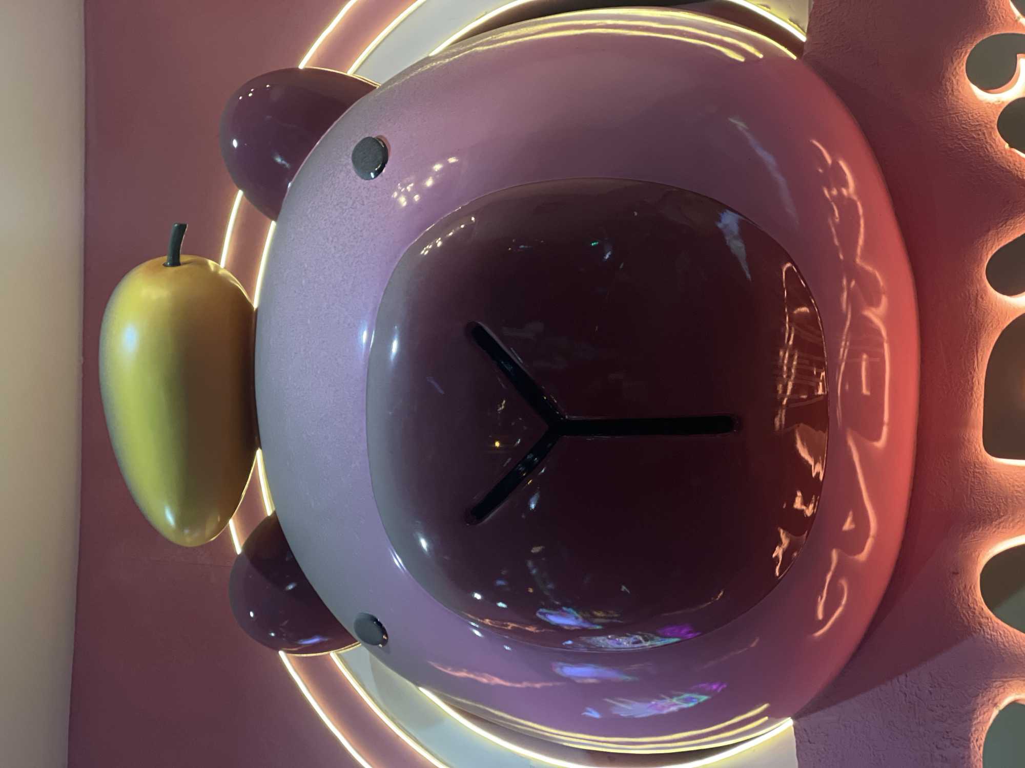 The image depicts a stylized, oversized pink bear head against a pink wall. A yellow fruit, possibly a mango, is positioned to the left of the head. The bear head is a rounded structure with a dark brown, shiny facial area featuring a stylized nose and mouth. The bear has round ears with black dots. The bear is surrounded by a circular, neon-lit ring. A textured pink element with holes is positioned at the bear's right side. The photograph was taken in Nha Trang Ward, Vietnam.