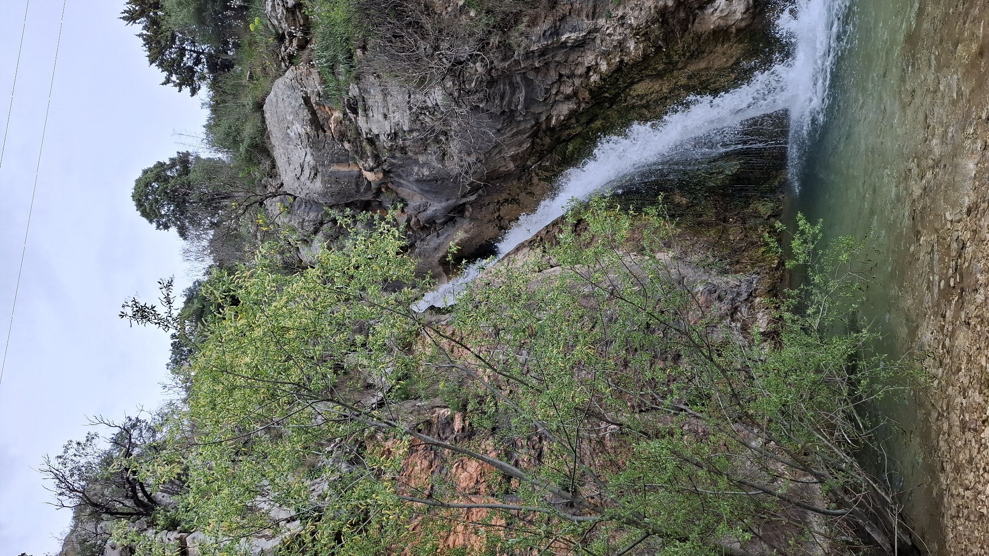The image depicts a waterfall in the city of Boreti, Montenegro. The water cascades from a rocky cliff into a small pool. The cliff face is a mix of gray and reddish-brown rock. Vegetation grows on and around the rocks, including green trees and shrubs. The water in the pool has a greenish tint. The ground surrounding the pool is covered in small stones. The sky is overcast.