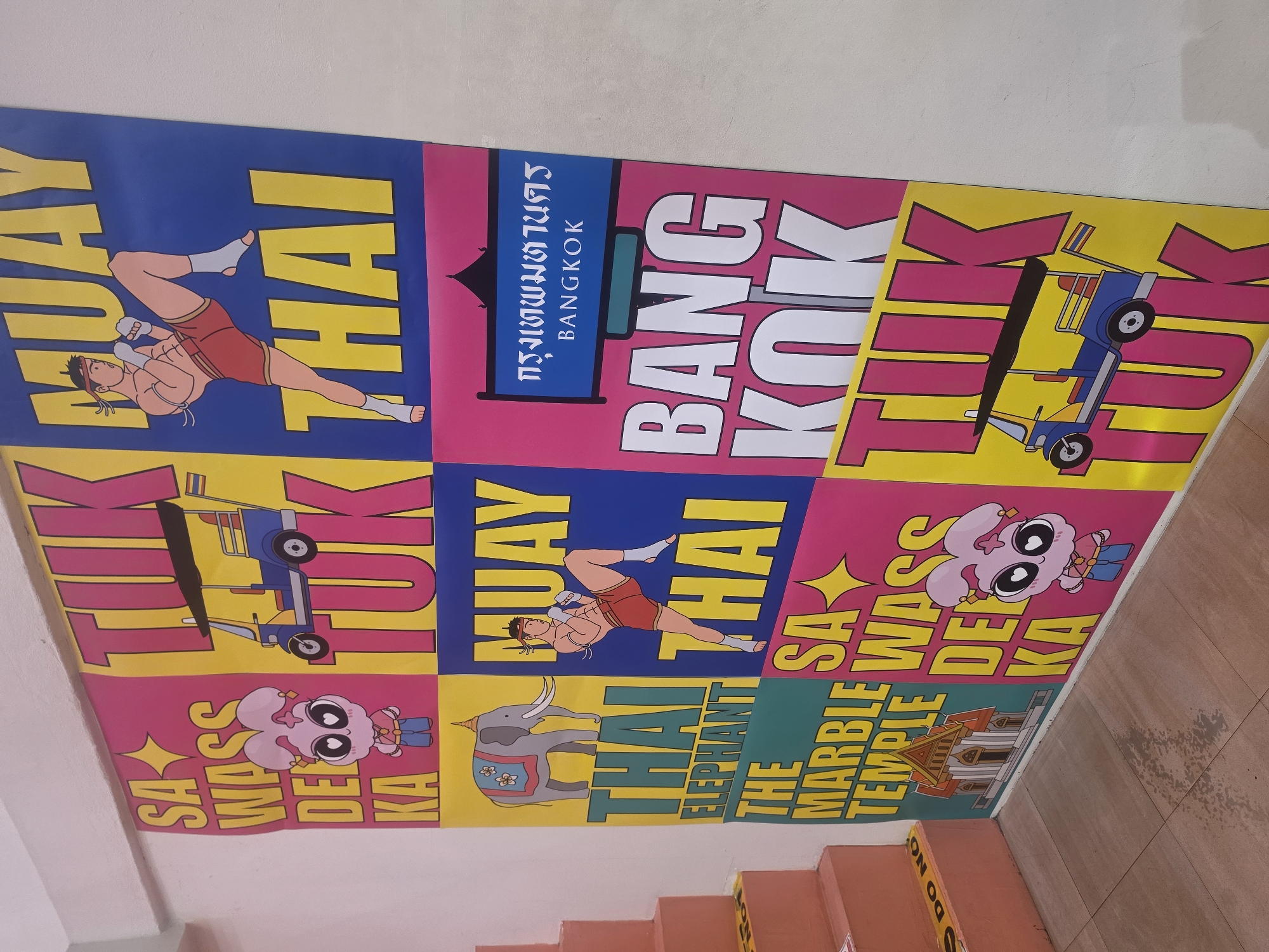 This is a vibrant and colorful mural, likely on a wall or ceiling, showcasing various iconic elements of Thai culture and Bangkok. The mural is divided into multiple square or rectangular panels, each featuring a distinct illustration and accompanying text.
Subjects and objects depicted include:
* A male Muay Thai fighter in a dynamic pose, featured in two different blue panels.
* A traditional Thai Tuk-Tuk vehicle, depicted in two separate yellow and pink panels.
* A large gray elephant with decorative elements, shown in a yellow panel.
* A cartoonish pink creature with large eyes, appearing in two pink panels.
* A stylized depiction of a temple building, specifically "The Marble Temple," in a green panel.
The overall scene functions as a visual celebration or advertisement for Bangkok, featuring popular symbols and attractions. The bright, saturated colors of blue, pink, yellow, and green, along with the cartoonish art style, give the mural a modern, pop-art aesthetic.
Visible text within the mural includes:
* "MUAY THAI" (appearing twice)
* "กรุงเทพมหานคร" (Krung Thep Maha Nakhon, meaning Bangkok in Thai script)
* "BANGKOK"
* "TUK TUK" (appearing twice)
* "SA WASS DE KA" (a common Thai greeting, appearing twice)
* "THAI ELEPHANT"
* "THE MARBLE TEMPLE"
The image also shows a light-colored wall or ceiling surrounding the mural, and hints of red-orange steps are visible at the bottom, suggesting the mural might be in a stairwell or public space. The image itself is rotated, but the crops clarify the content.