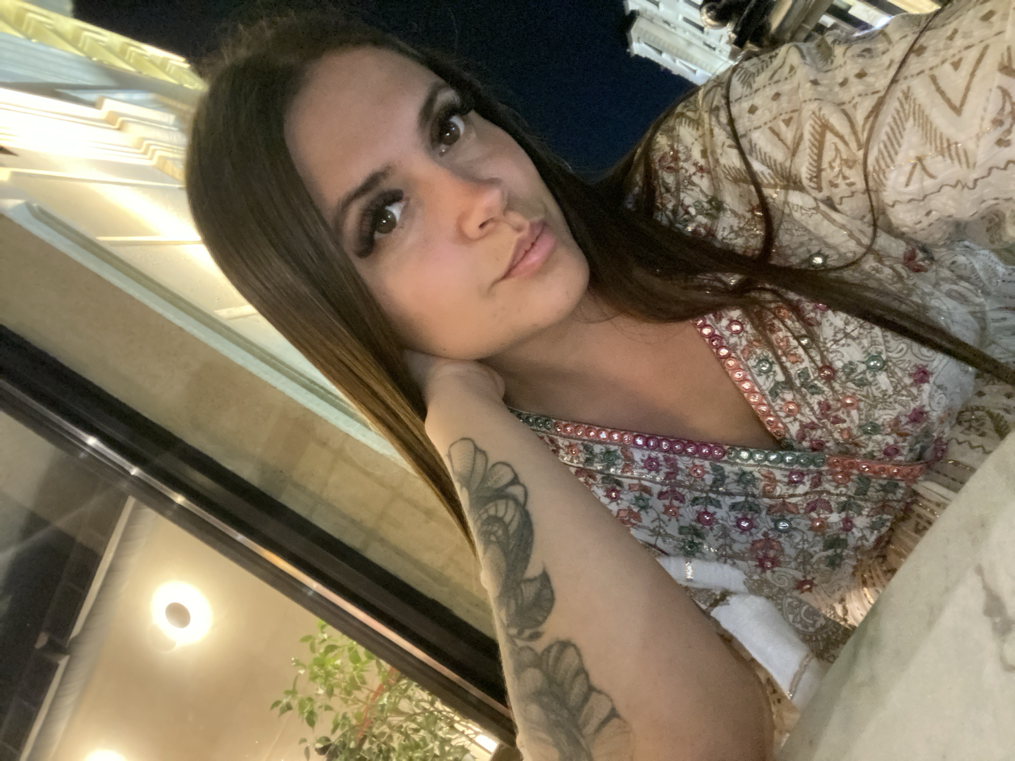 In an outdoor urban setting at night, a young woman with long, dark brown hair is seen from the chest up, looking towards the camera. She is resting her left arm on what appears to be a table in the foreground, with her hand positioned near her neck. She is wearing a white garment adorned with colorful floral embroidery and shimmering sequins. A tattoo is visible on her left arm. Behind her, out of focus, is a building with architectural details and illuminated windows, suggesting a restaurant or establishment. The background also shows a reflection in a window, hinting at an indoor space with lights and plants. The lighting suggests artificial sources, contributing to a nighttime ambiance. The specific location in Derbent, Russia, cannot be determined from the visual information alone.
