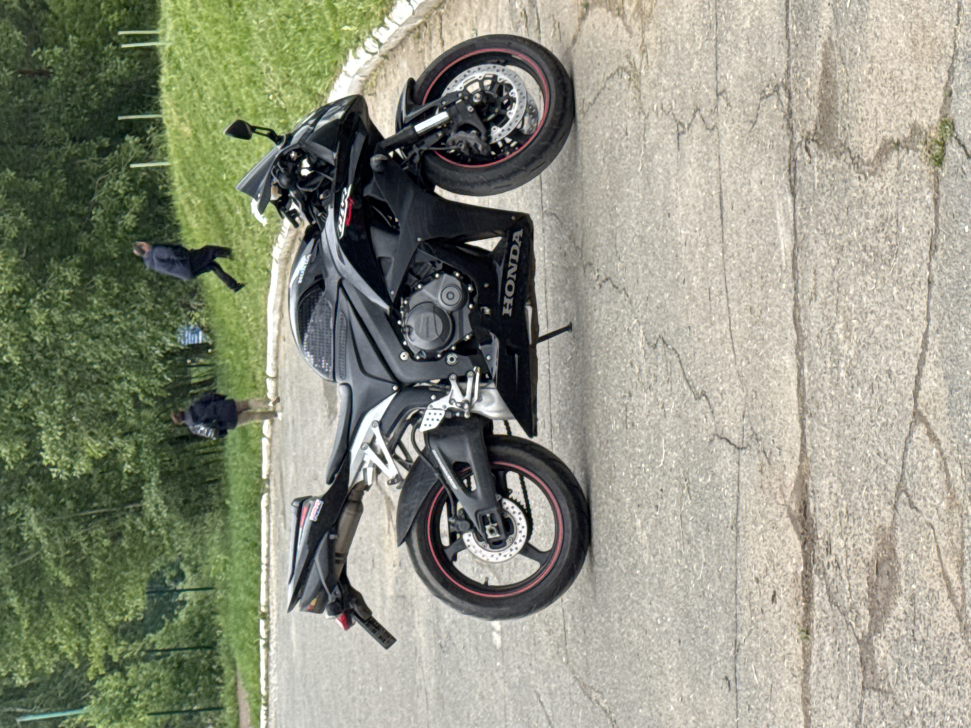 The image shows a black Honda motorcycle parked on an asphalt surface with visible cracks. "HONDA" is printed on the motorcycle's fairing. In the background, there's a grassy area with trees, and two individuals are walking in the distance. The location is likely Chaikovsky, Russia.