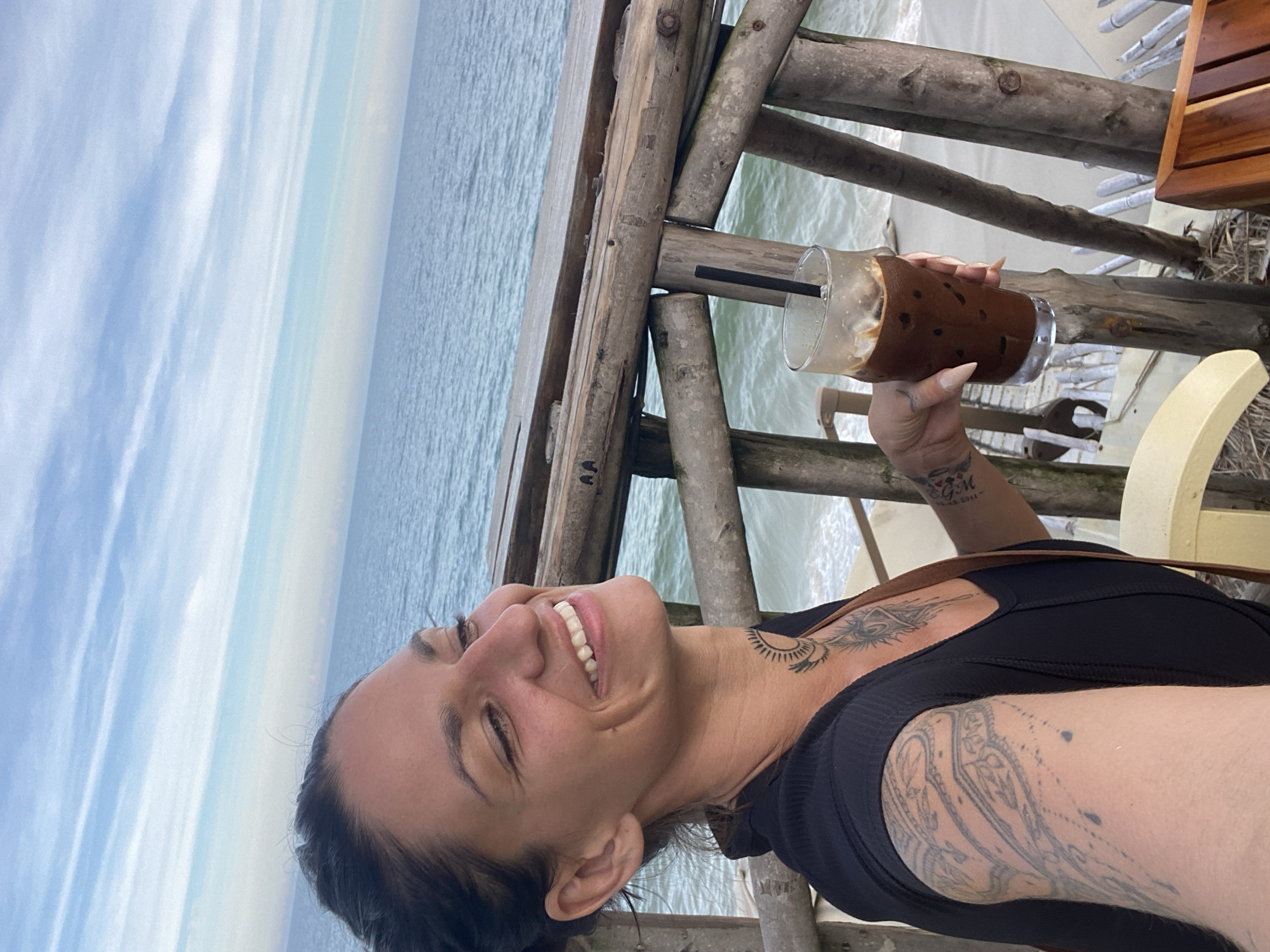 A woman with dark hair pulled back and a black top smiles while holding an iced coffee. She has tattoos on her arm and neck. She is seated at a table on a wooden deck overlooking the ocean. The sky is blue with white clouds. The image is taken from a high angle looking down at the woman. The location is Phu Thuy Ward, Vietnam.