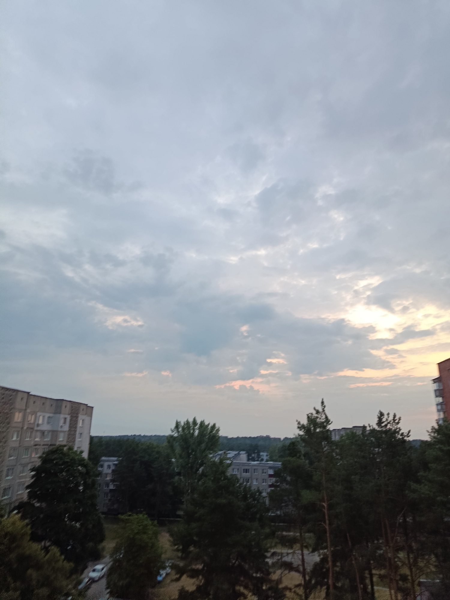 This is a high-angle, eye-level shot taken from a residential building in Svietlahorsk, Belarus, looking towards the west over a mixed urban and natural landscape under a cloudy sky. The sky is dominant, featuring a mix of grey and white clouds with areas of brighter, possibly sunset-lit, sky peeking through on the right side. In the foreground, silhouetted pine trees are visible, some with distinct vertical trunks. Behind the trees, a cluster of multi-story residential buildings are present, some appearing to be older Soviet-era apartment blocks. One building on the left is light-colored and has many windows. Further back, more residential structures and dense green trees form a treeline across the middle ground. The lower portion of the image shows a street with at least two parked cars, one white and one blue, on the left side. The overall scene suggests a view from a balcony or window in a suburban or residential area of the city.